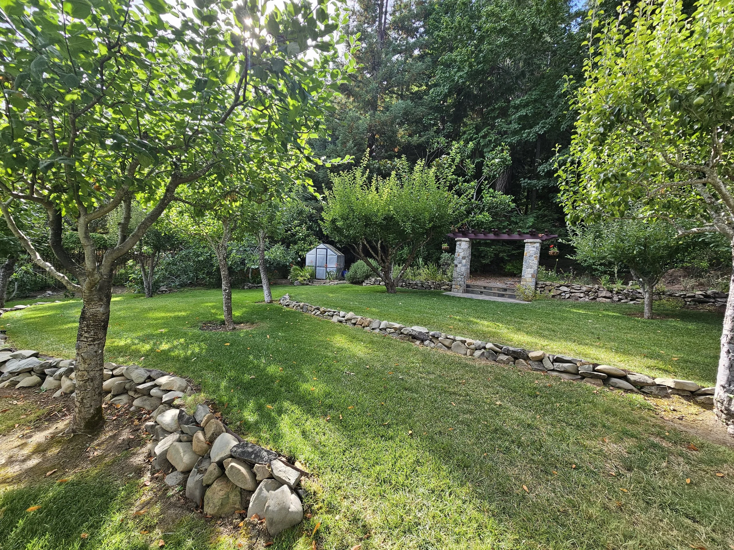 A lush backyard garden with several trees, a green greenhouse in the background, and a stone pergola. The area is well-maintained with a grassy lawn and a winding rock-lined border.