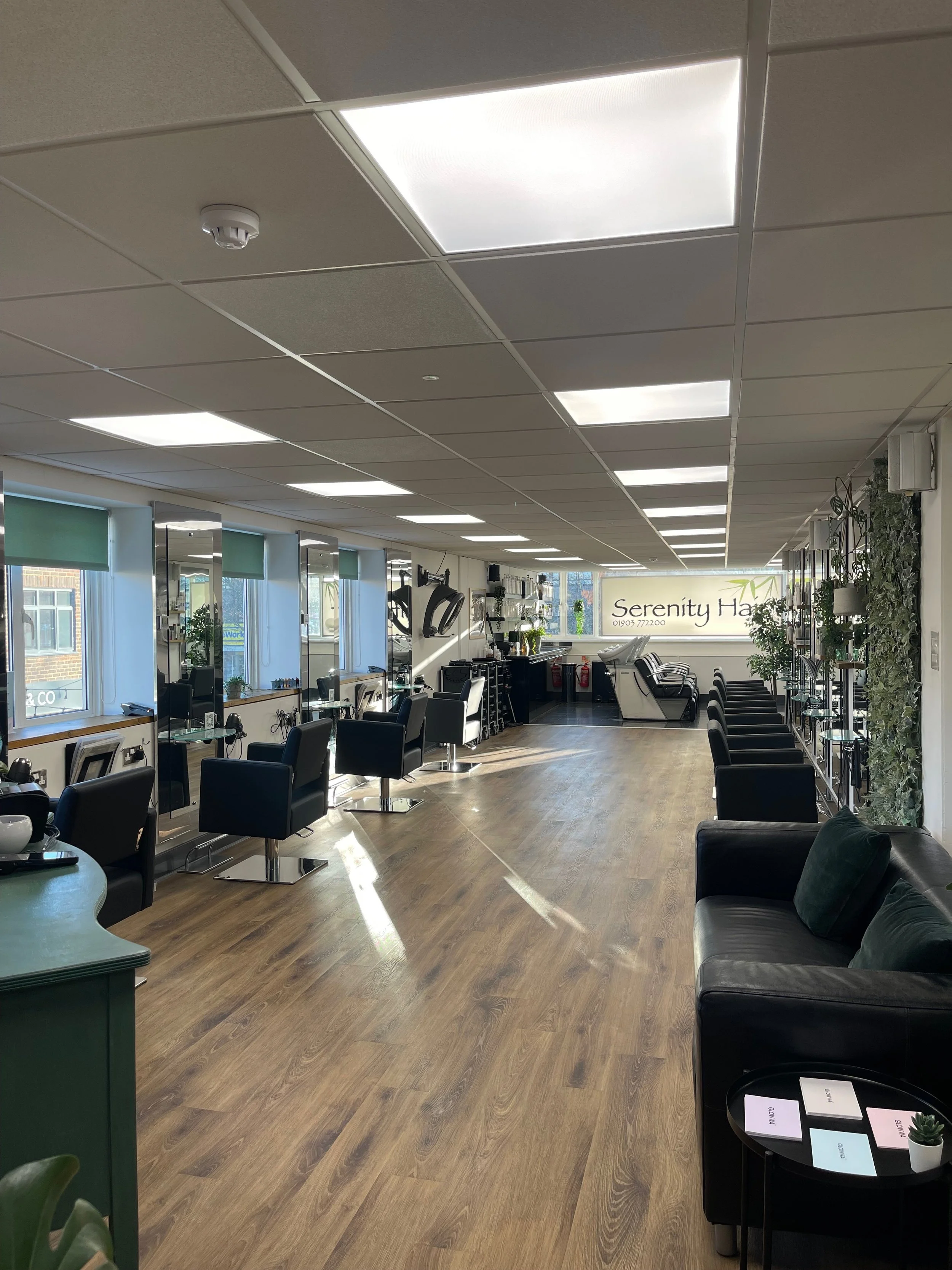 Empty hair salon with multiple styling stations, black chairs, mirrors, and a waiting area with black sofa and small table, illuminated by overhead lighting.