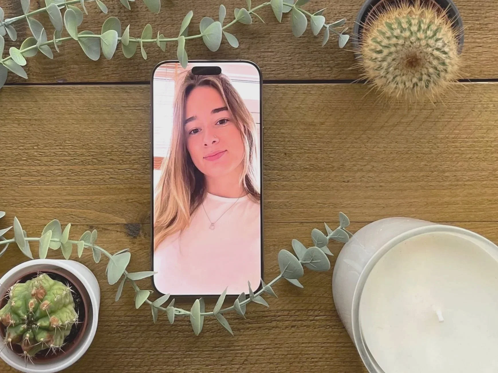 A smartphone on a wooden surface displaying a photo of a smiling young woman with long, wavy, blonde hair and a white top. Surrounding the phone are potted cacti, a small potted plant with green leaves, and a white candle in a round container.