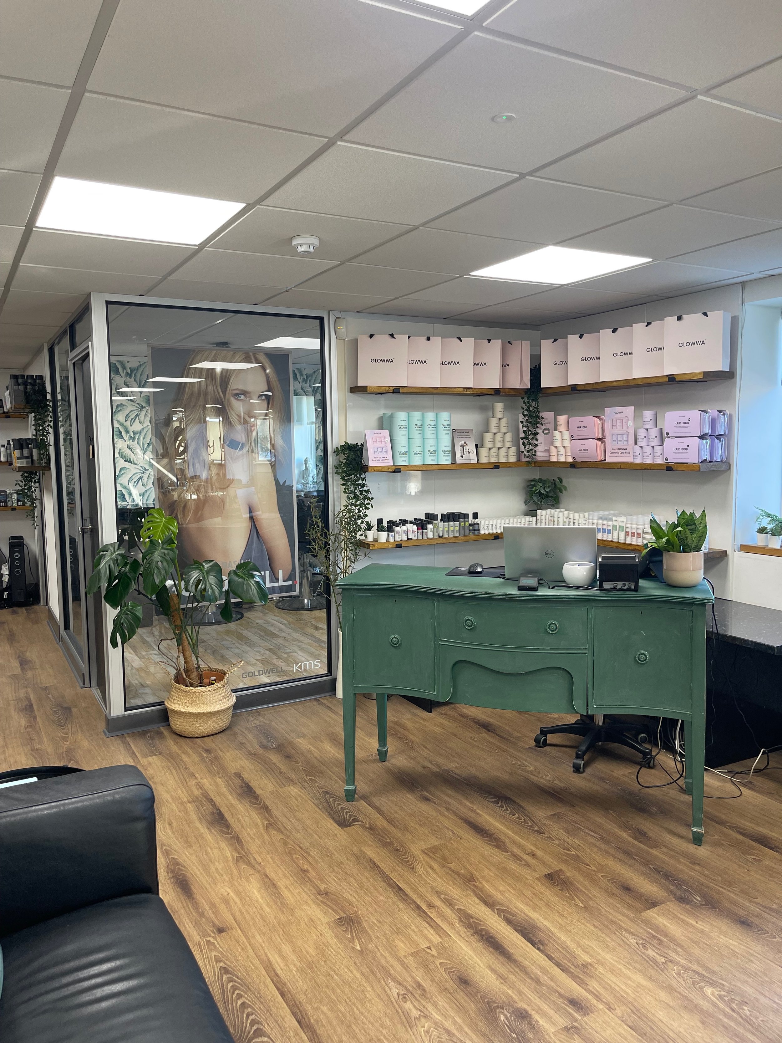 Hair salon with green reception desk and black sofa with retail products on the shelves, decorated with plants.
