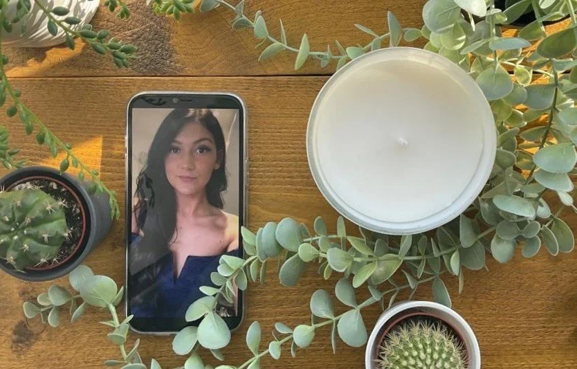 A wooden table with a smartphone displaying a woman's photo, surrounded by potted succulents and cacti, and a large white candle.