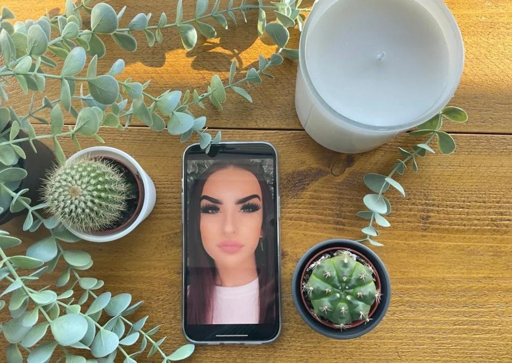 A smartphone displaying a woman with makeup, surrounded by potted plants and a lit candle on a wooden surface.
