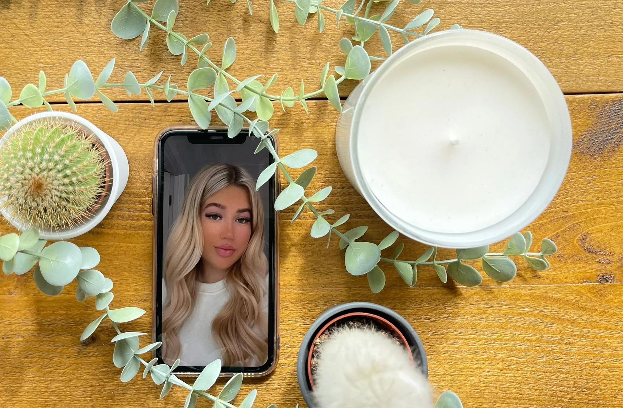 A smartphone laying on a wooden surface, displaying a woman's face with blonde hair. Surrounding the phone are potted plants, including a cactus and eucalyptus leaves, and a large white candle.