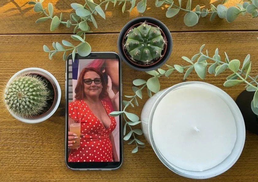 A wooden table with a smartphone displaying a woman in a red polka dot dress holding a drink, surrounded by cactus and eucalyptus plants, and a large white candle.