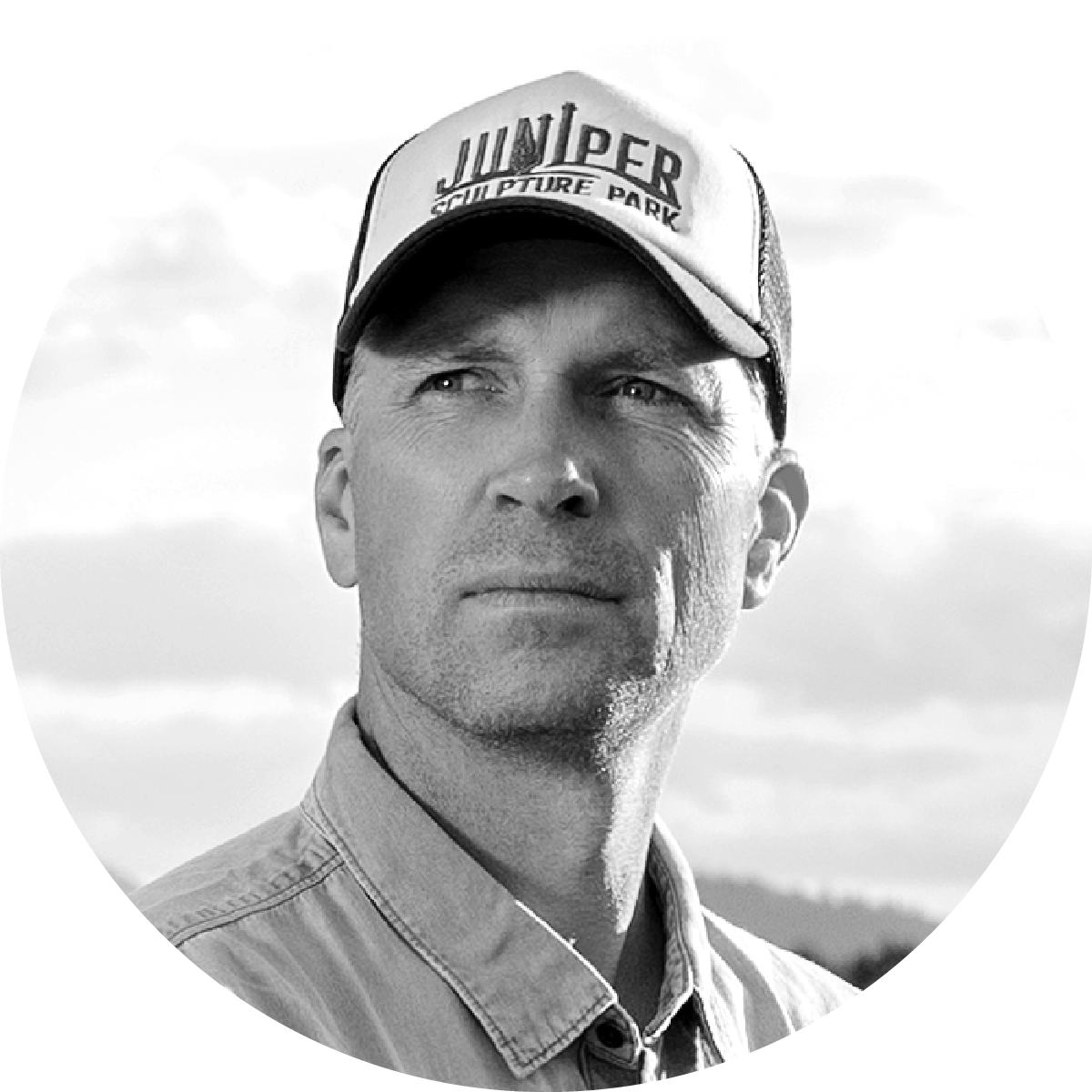 Black and white photo of a man wearing a cap with "JUNIPER SCULPTURE PARK" written on it, looking into the distance with a serious expression, outdoors with a cloudy sky in the background.