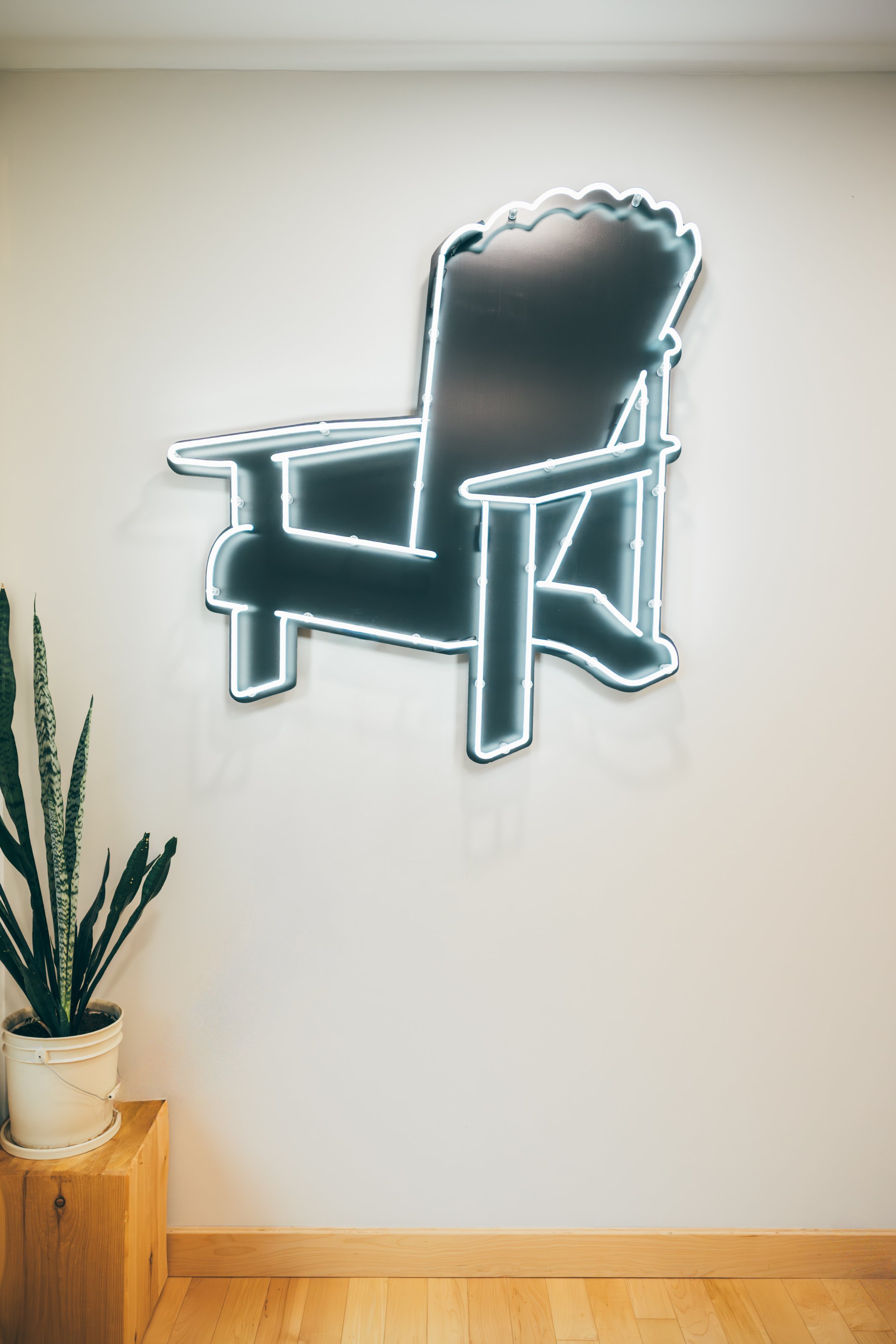 A neon sign shaped like high back Adirondack style chair on a plain white wall, with a potted snake plant on a wooden stand in the corner.