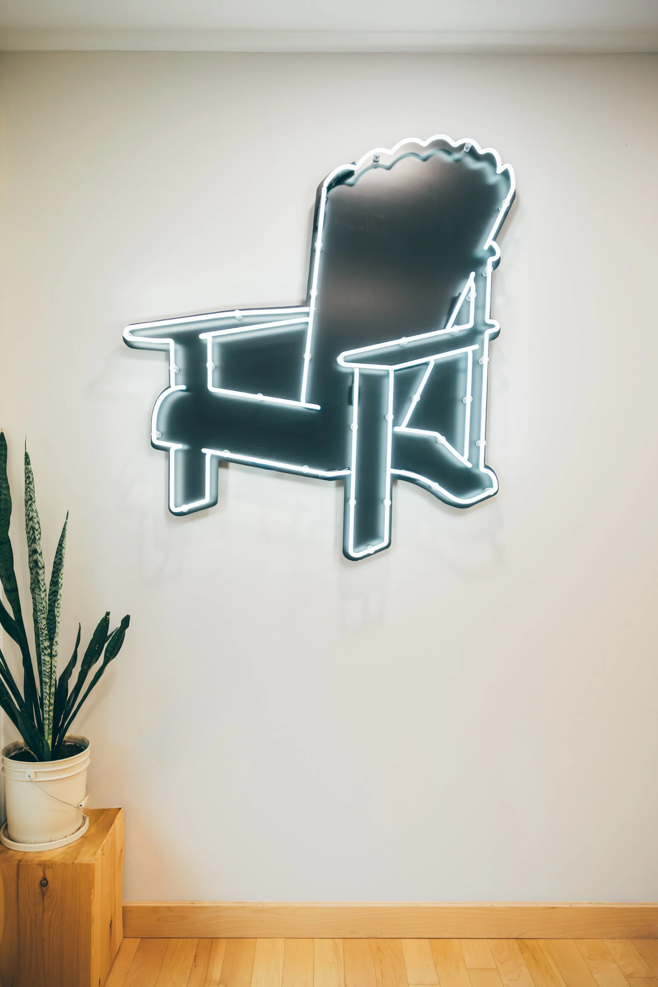 A neon sign shaped like high back Adirondack style chair on a plain white wall, with a potted snake plant on a wooden stand in the corner.