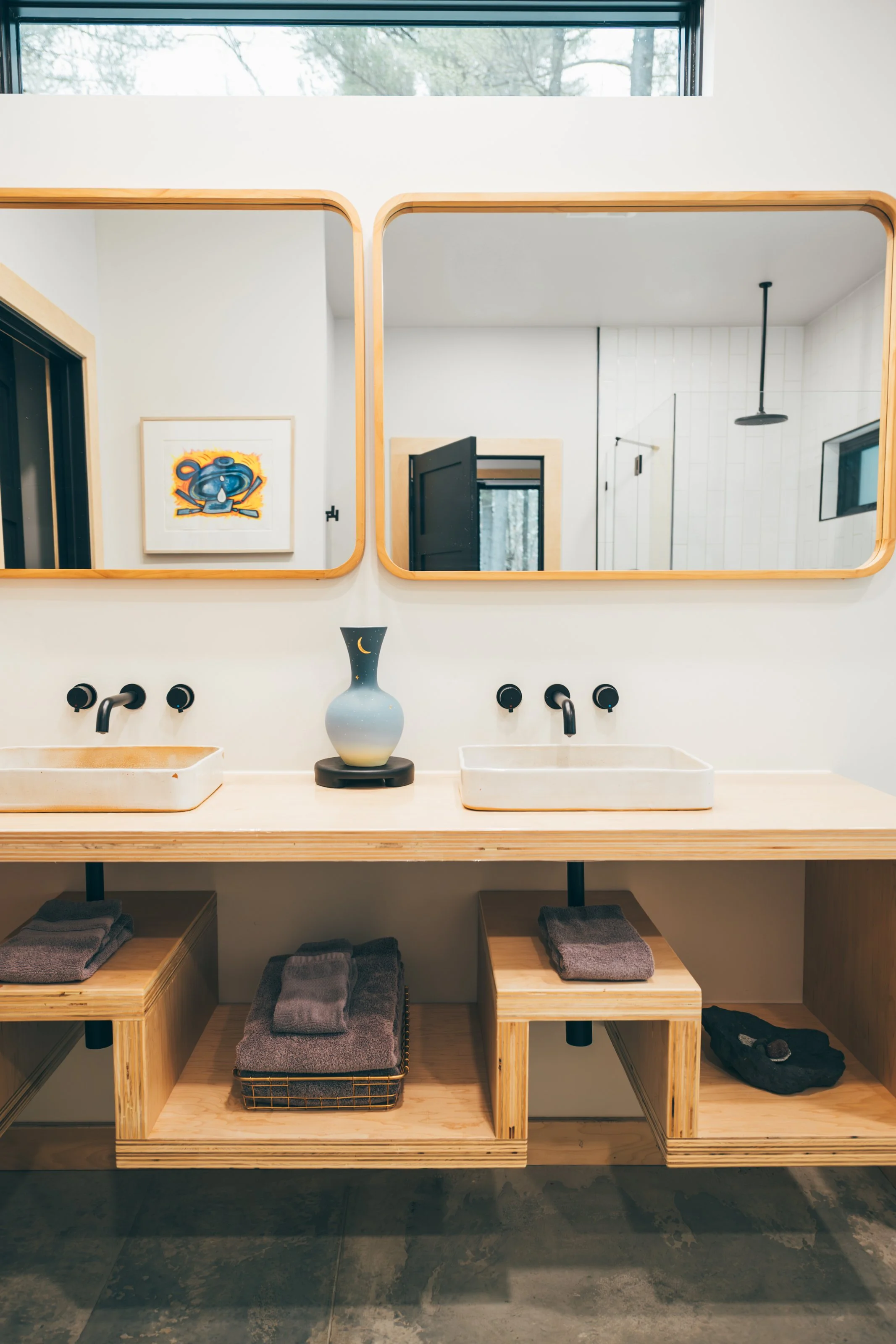 Wooden bathroom vanity with two vessel sinks, black wall-mounted faucets, a blue and beige vase, and neatly folded gray towels on open shelves beneath. There are two wooden framed mirrors above the sinks reflecting part of a modern interior and a pie