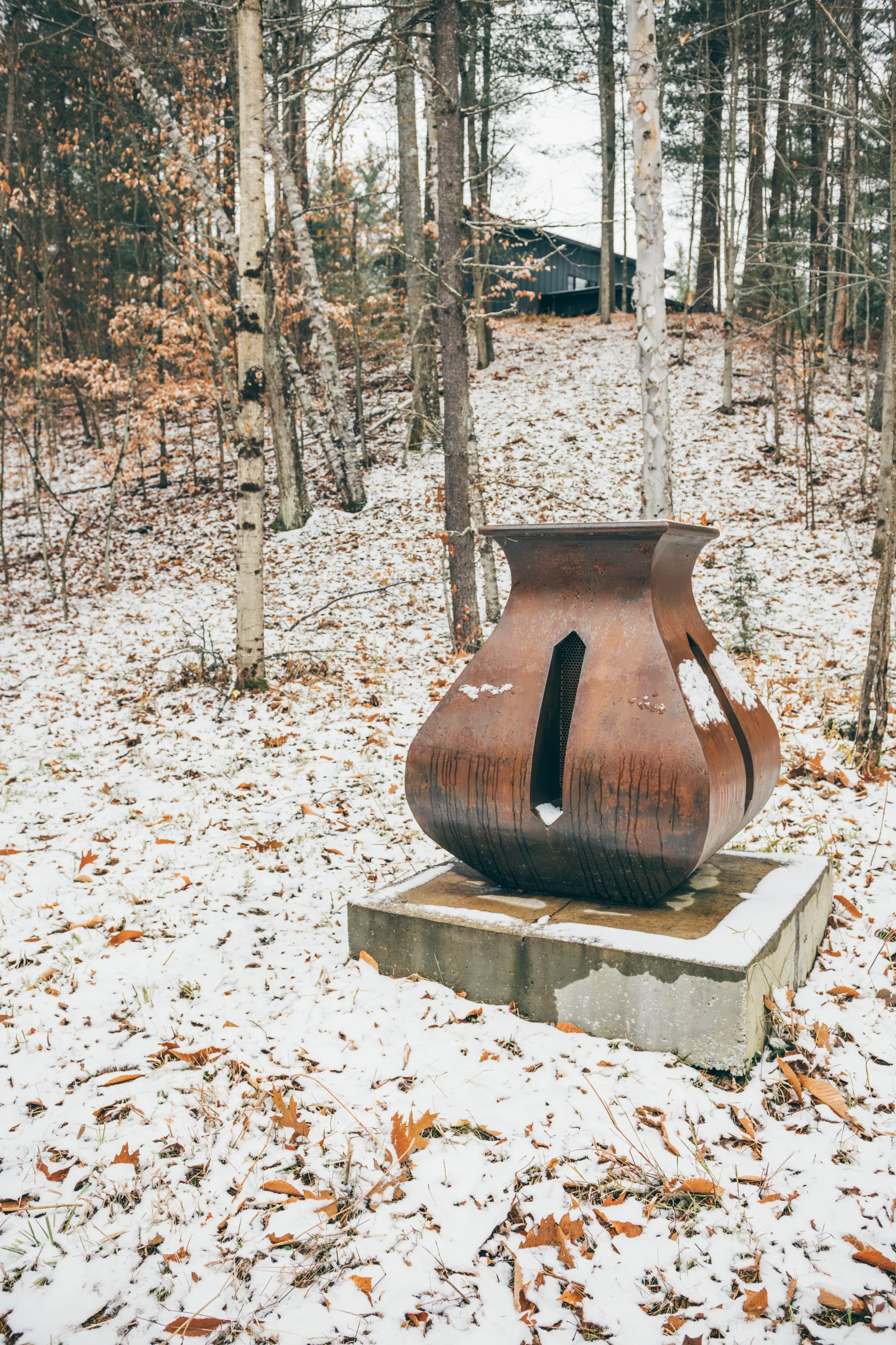 Rusty outdoor sculpture on concrete base in a snowy wooded area with a house in the background.