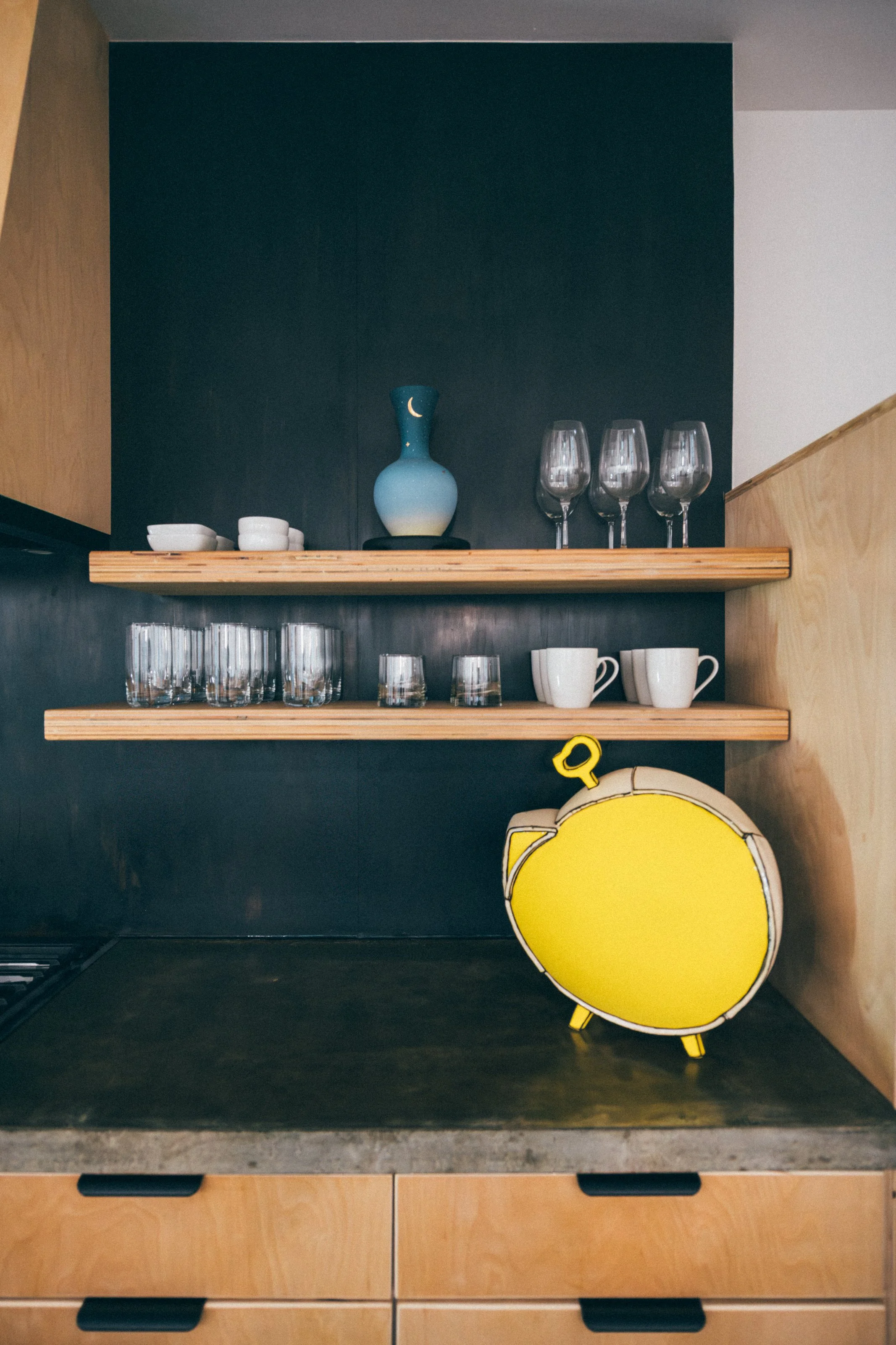 Kitchen shelf with white bowls, wine glasses, coffee mugs, and decorative ceramic vase with moon and star patterns, with a yellow sculpture in the forground.