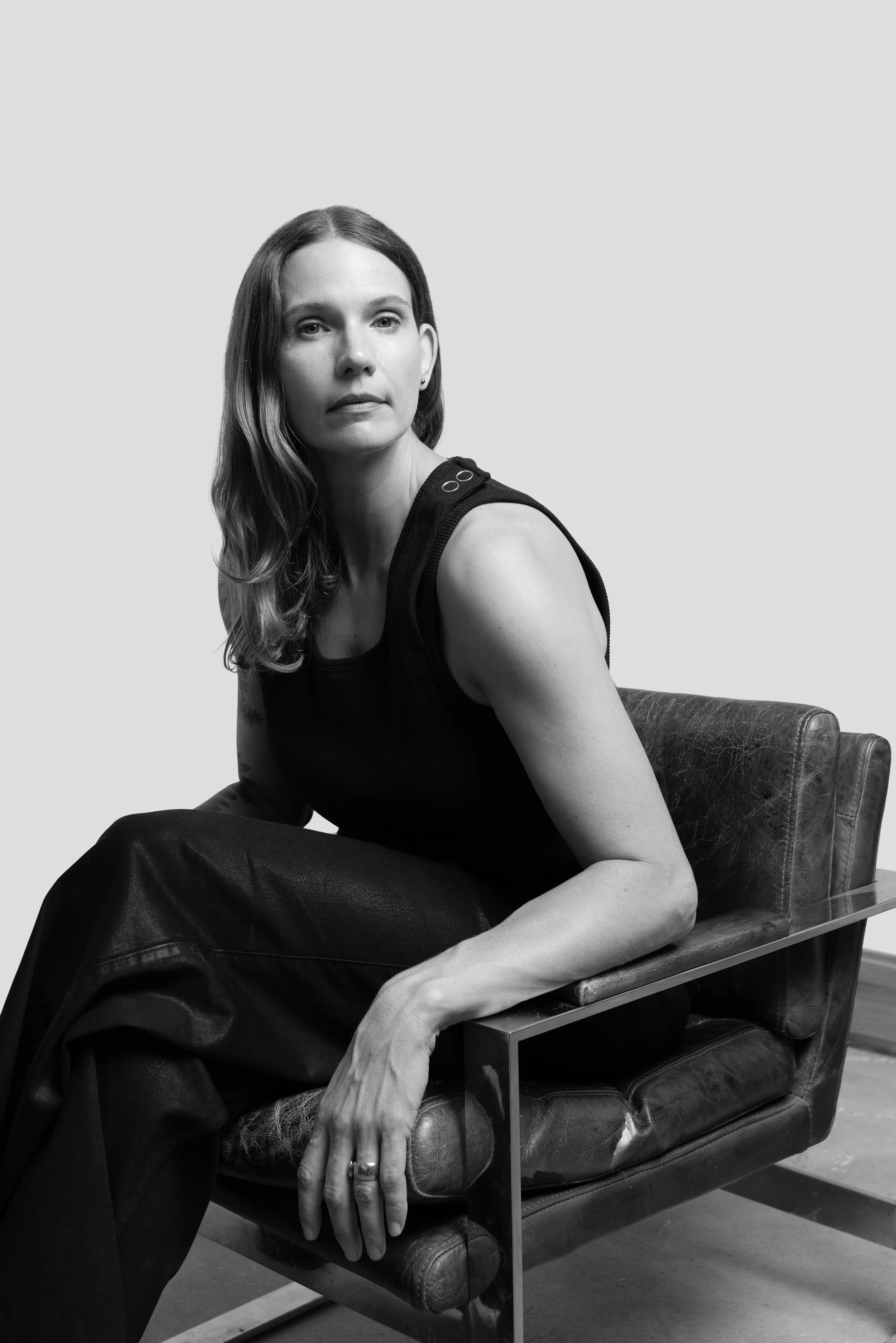 A woman with long wavy hair sitting on a vintage leather chair with metal framing, wearing a dark sleeveless top, in a black and white photo, looking toward the camera with a neutral expression.