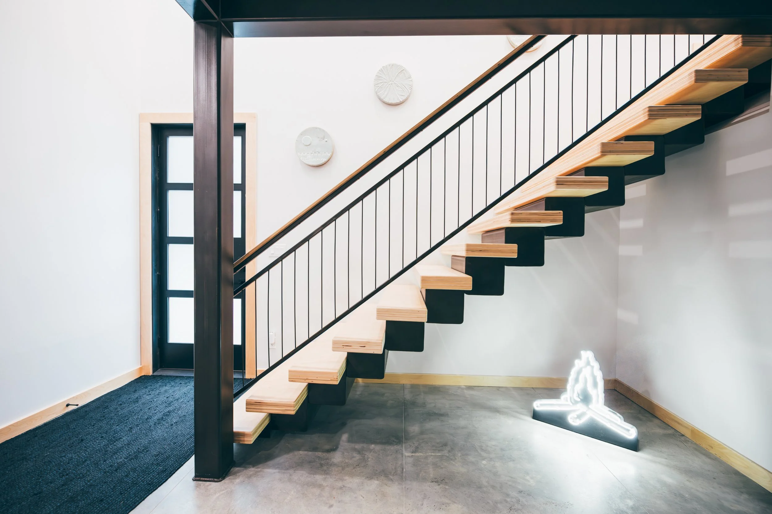 Modern staircase with open wooden steps and black supports, located in a bright room with white walls, a small window, and circular wall art. There is a neon light sculpture on the floor to the right.