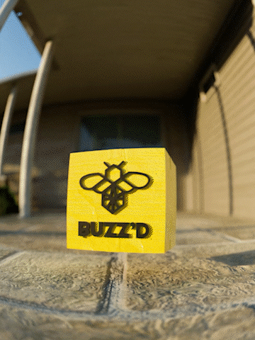 Yellow cube with a black bee logo and the word 'BUZZD' on it, placed on a stone surface outside a building.