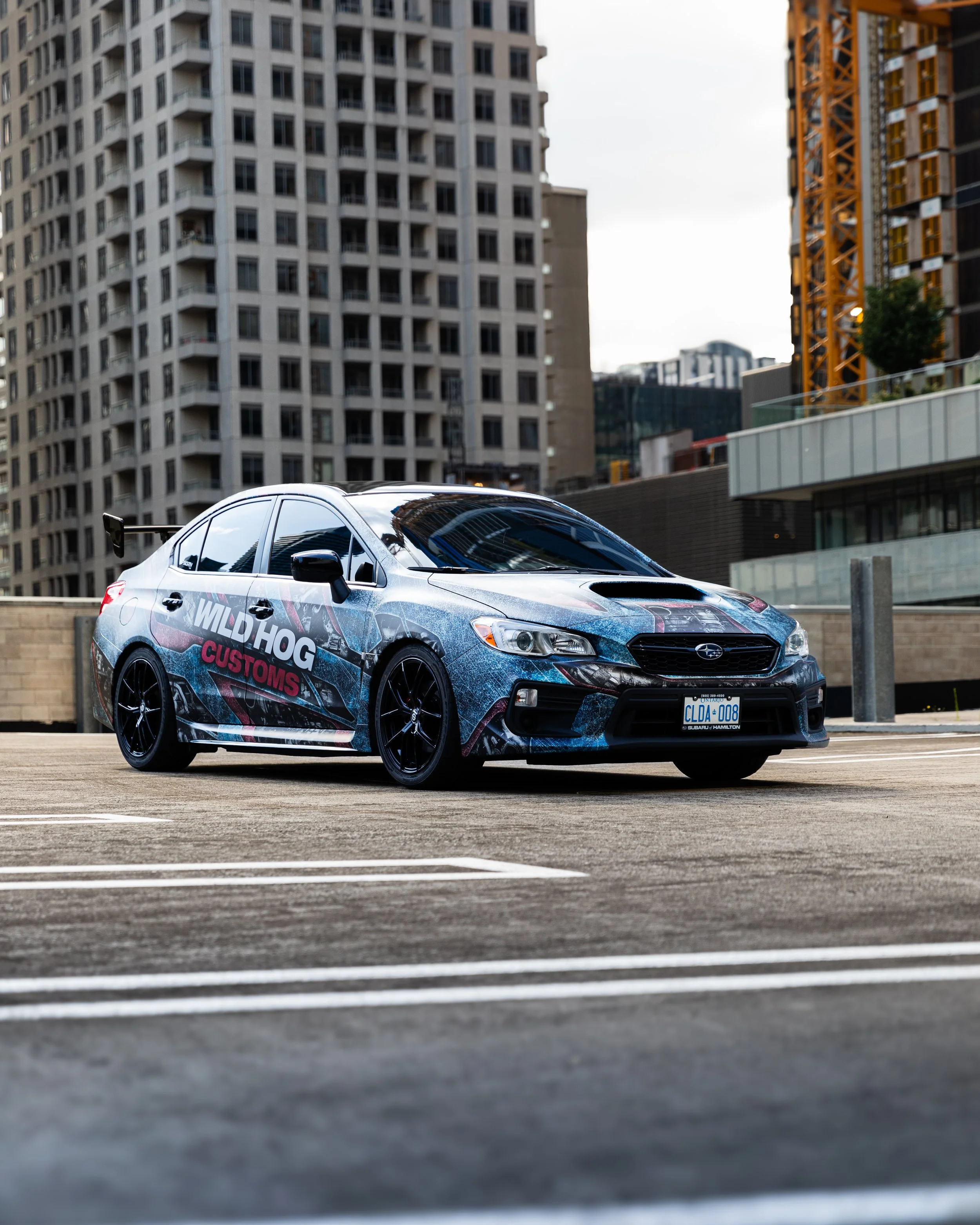 A black Subaru sedan with custom graphics and the words 'WILD HOG CUSTOMS' on the side, parked in an urban parking lot with tall buildings in the background.