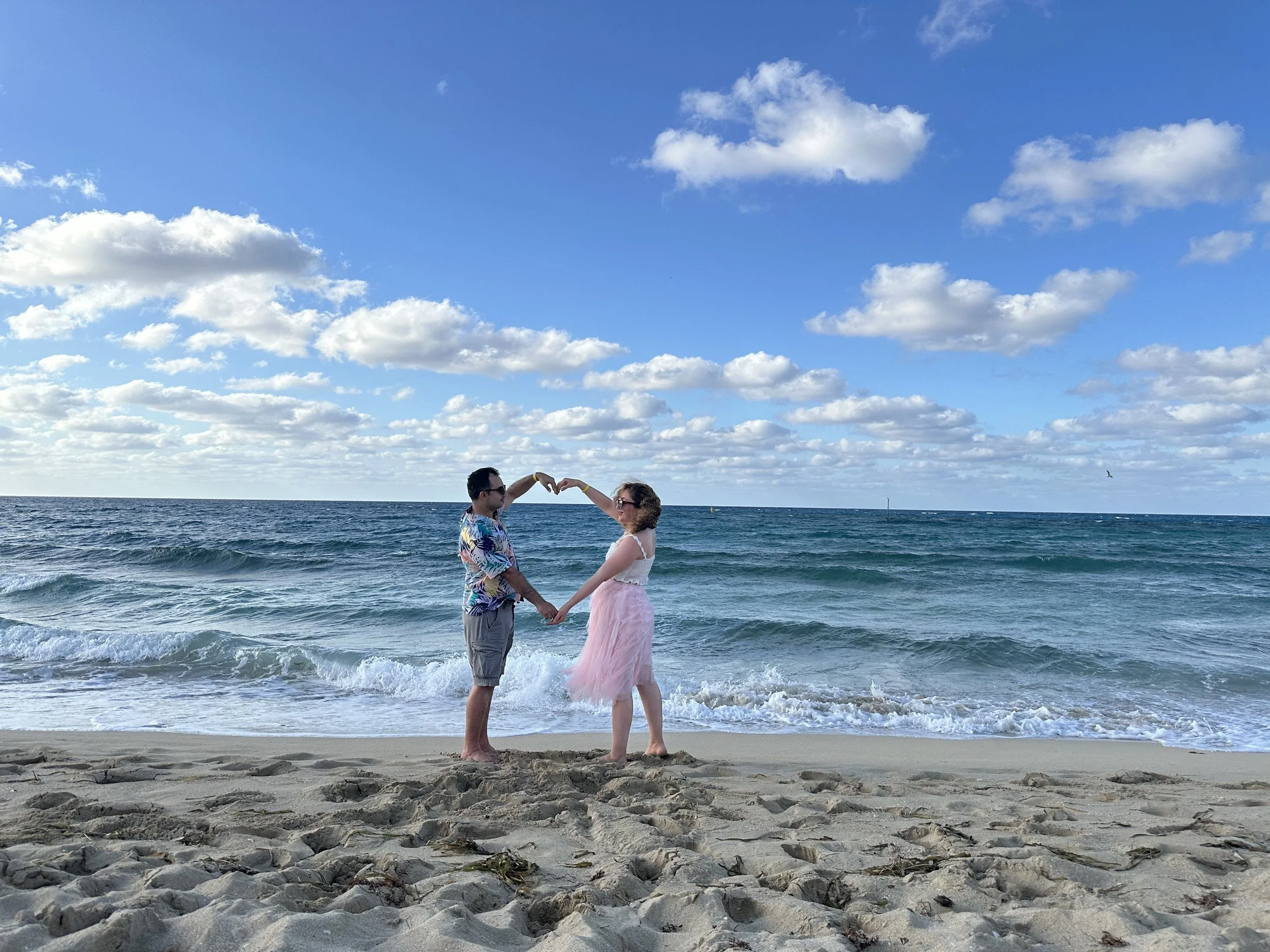 A couple holding hands on a sandy beach with the ocean and partly cloudy sky in the background, forming a heart shape with their arms.