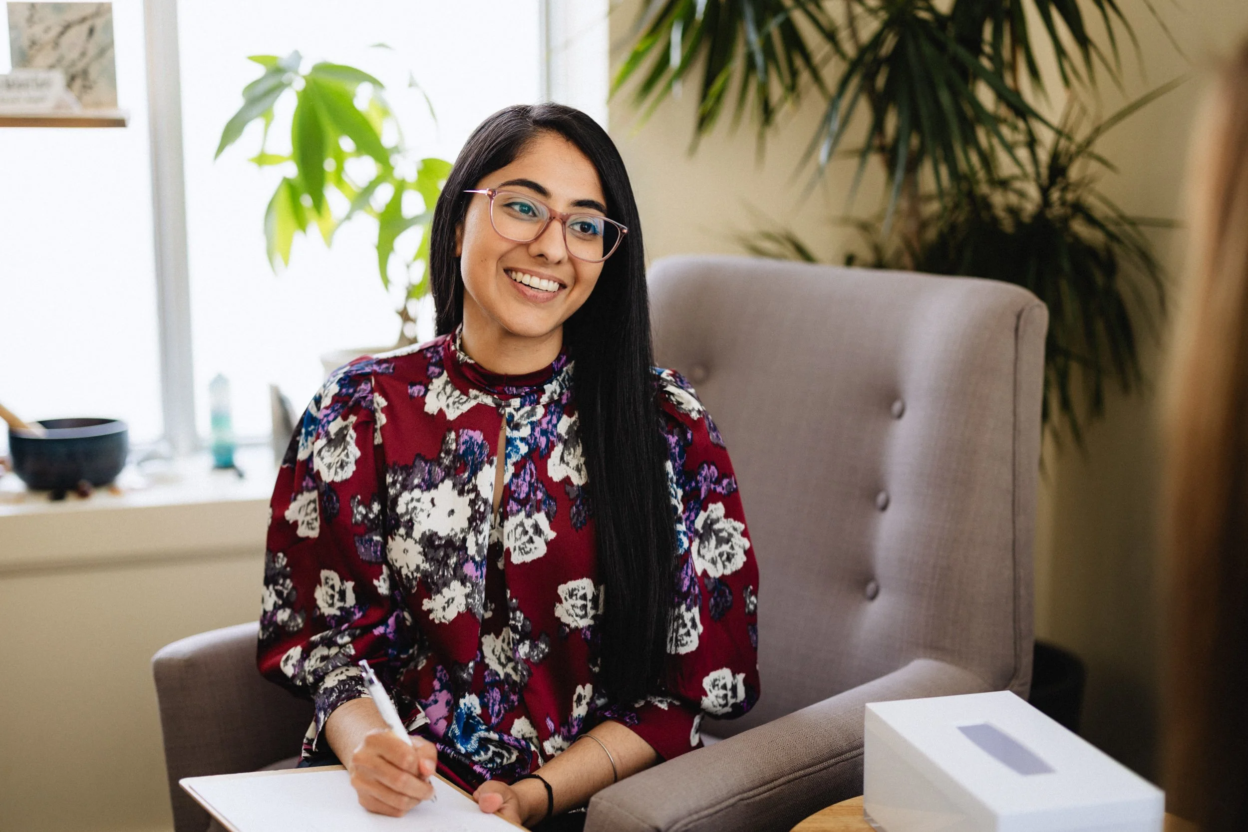 A woman with long black hair and glasses smiling while sitting in a room with a potted plant on the windowsill.