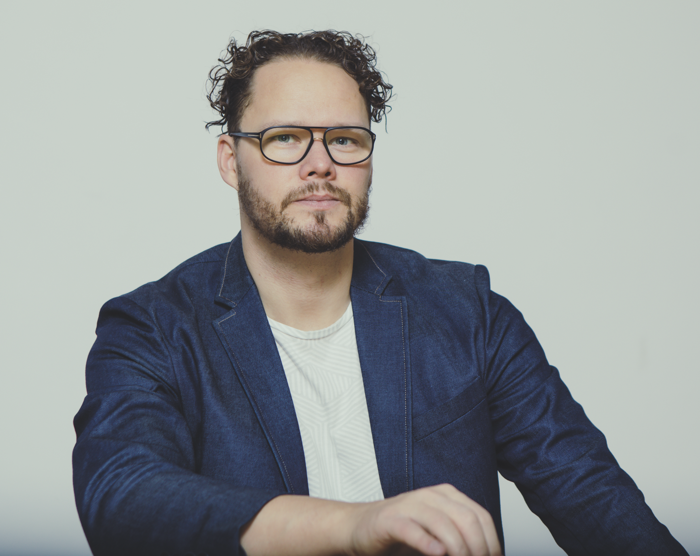 A man with curly blond hair, glasses, and a beard, wearing a blue blazer and white shirt, sitting with arms crossed against a plain light background.