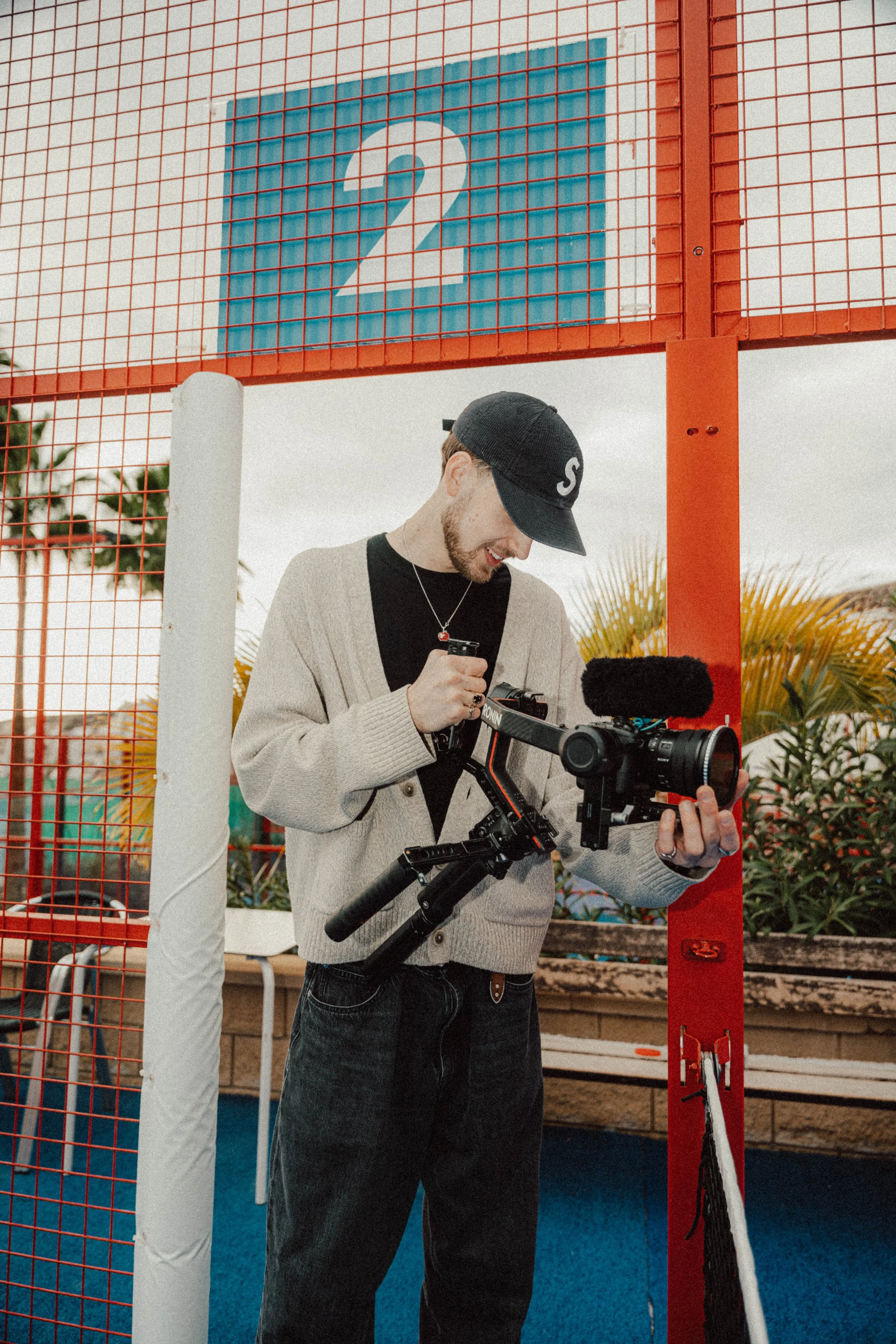 A man filming with a camera on a stabilizer at a sports court, with a sign featuring the number 2 behind him.