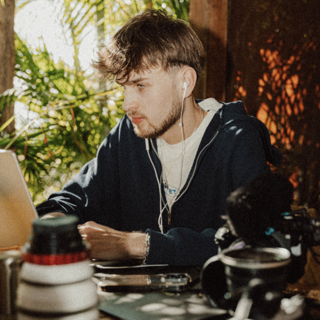A young man with brown hair and beard wearing a dark hoodie and white t-shirt, working on a laptop at a wooden table in a lush, sunlit outdoor setting.