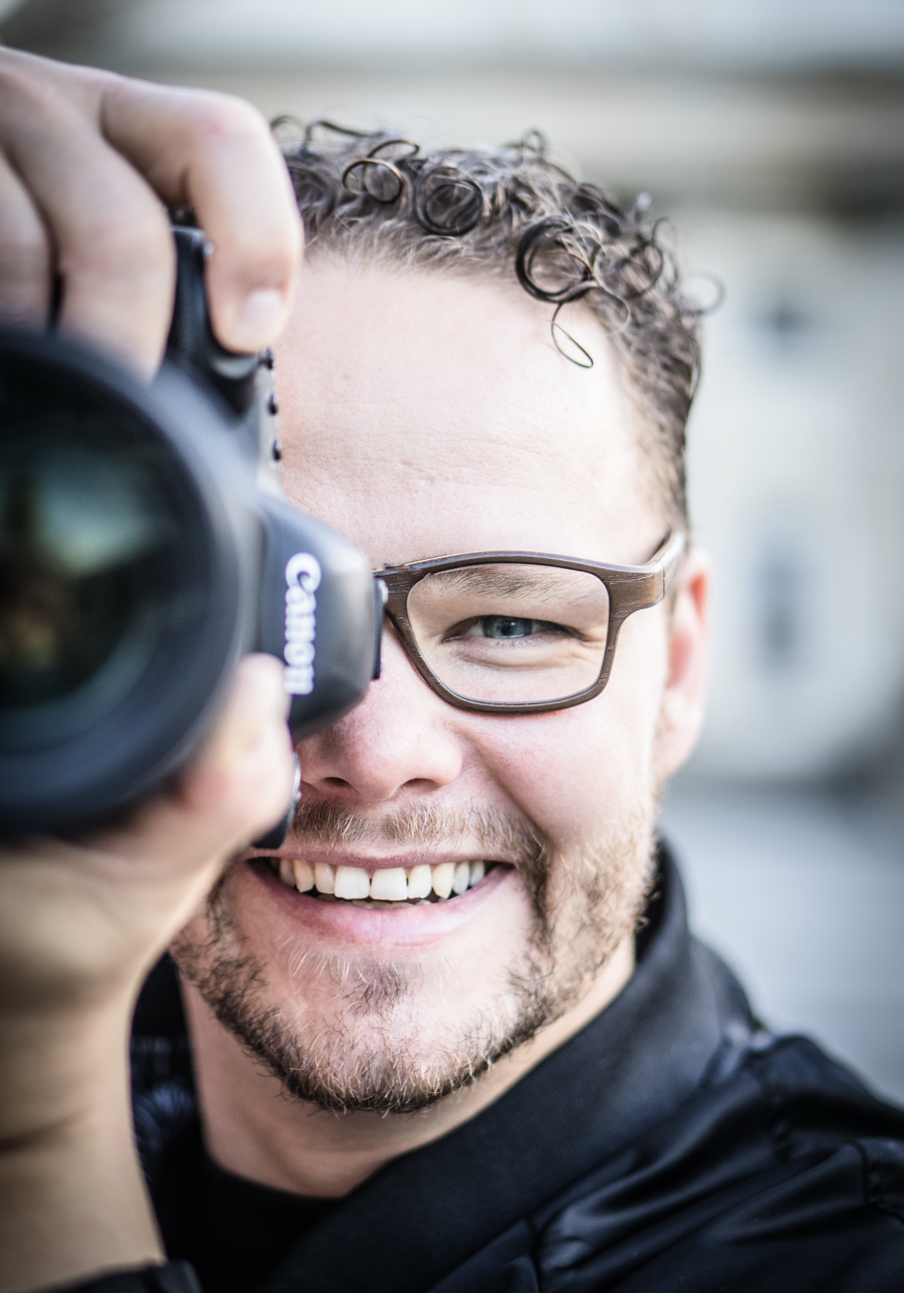A young man with curly hair and glasses smiling while taking a photo with a Canon camera.