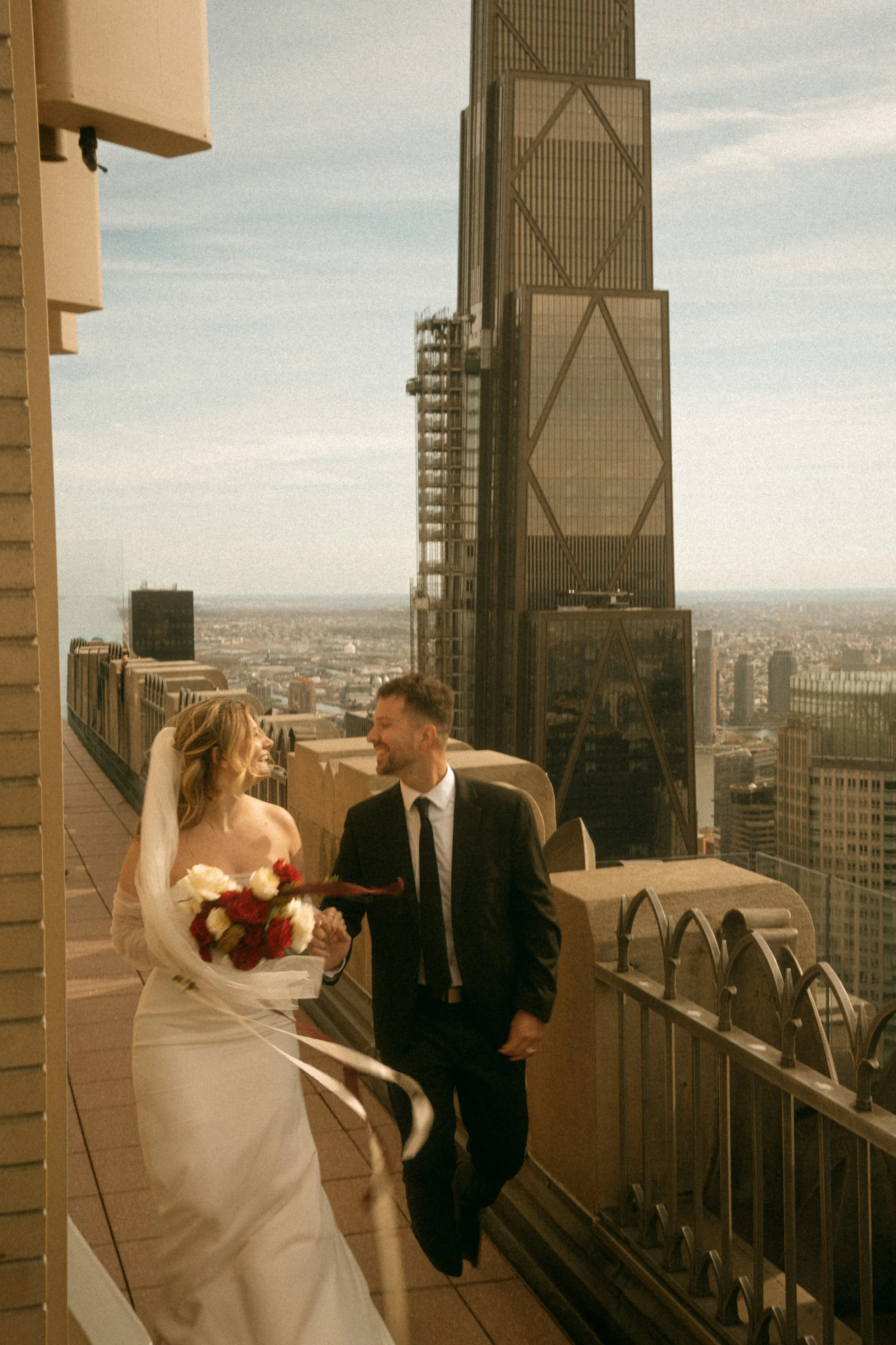 Tim & Emily at "Top of The Rock" at Rockefeller Center in NYC.   Emily holds a gorgeous red and white classic bouquet designed by New York City and Brooklyn florist, Raving Flamingo Flowers. Photo by Brittany Dobrowski, makeup by Lemony NYC.