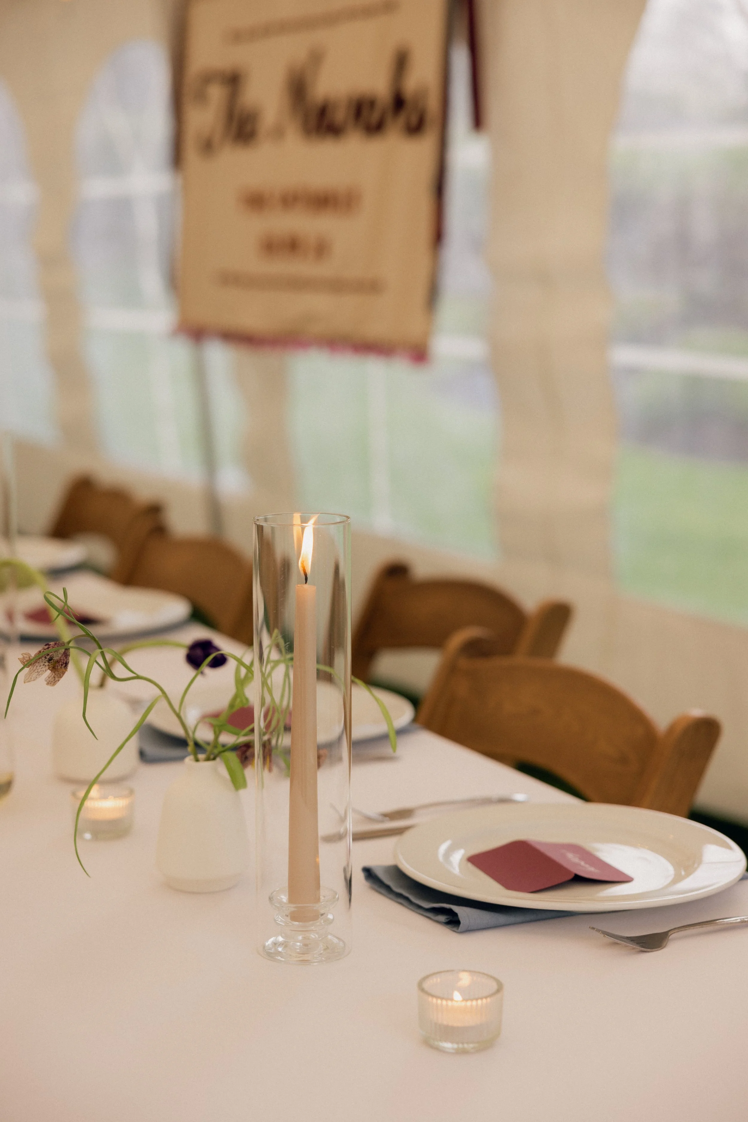 Elegant dining table set with a tall candle inside a glass holder and small votive candles, plates with pink napkins, silverware, a centerpiece with green vines and purple flowers, and a blurred background with a hanging wooden menu board and chairs 