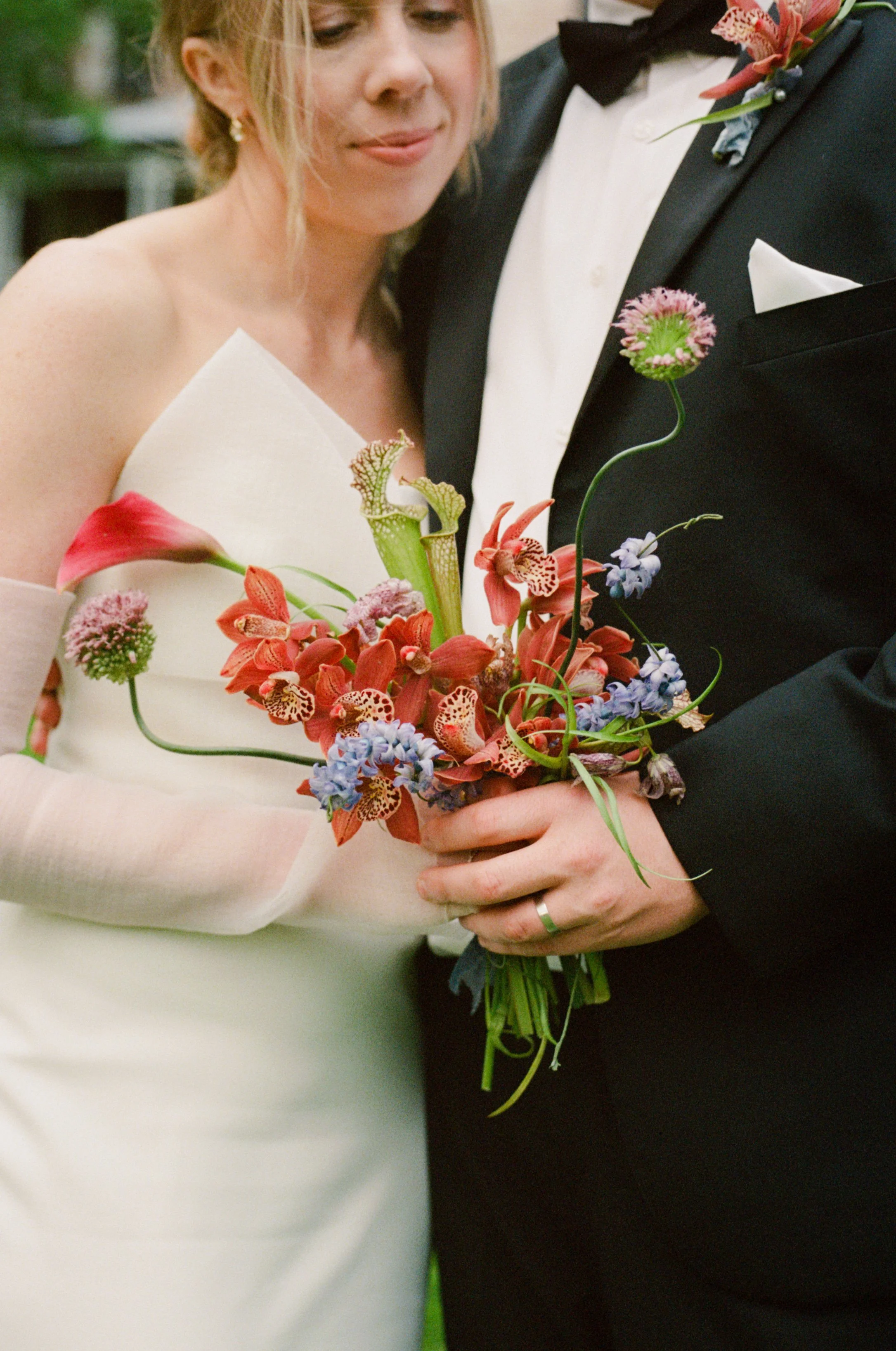Close-up of a bride and groom holding a bouquet of various flowers, with the bride wearing a white strapless wedding dress and the groom in a black tuxedo with a red boutonniere.