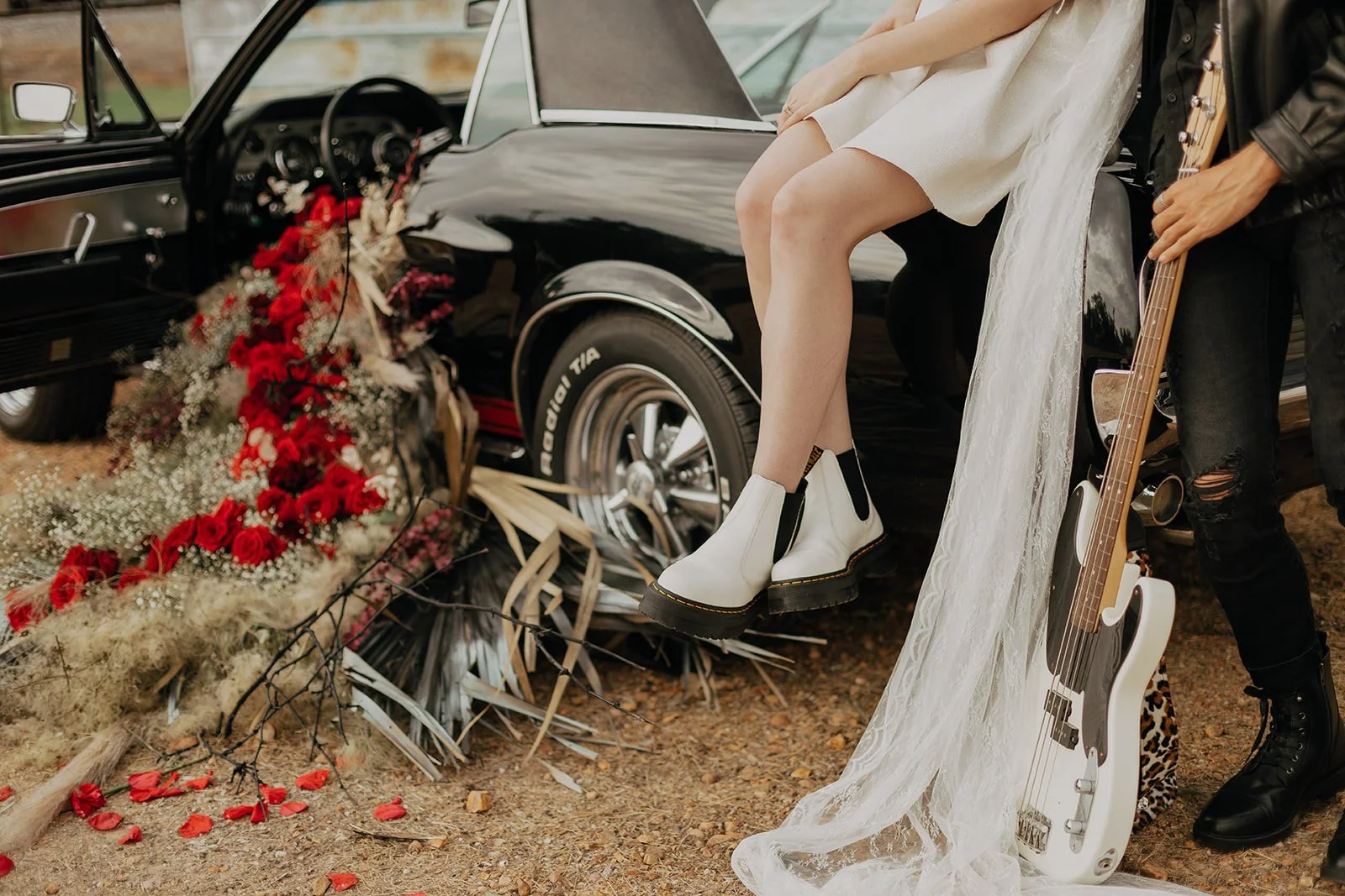 A woman in a white dress and white combat boots sitting on the edge of a black vintage car with a floral arrangement of red roses and white baby's breath on the ground nearby. A man in ripped black jeans and a leather jacket stands beside her holding