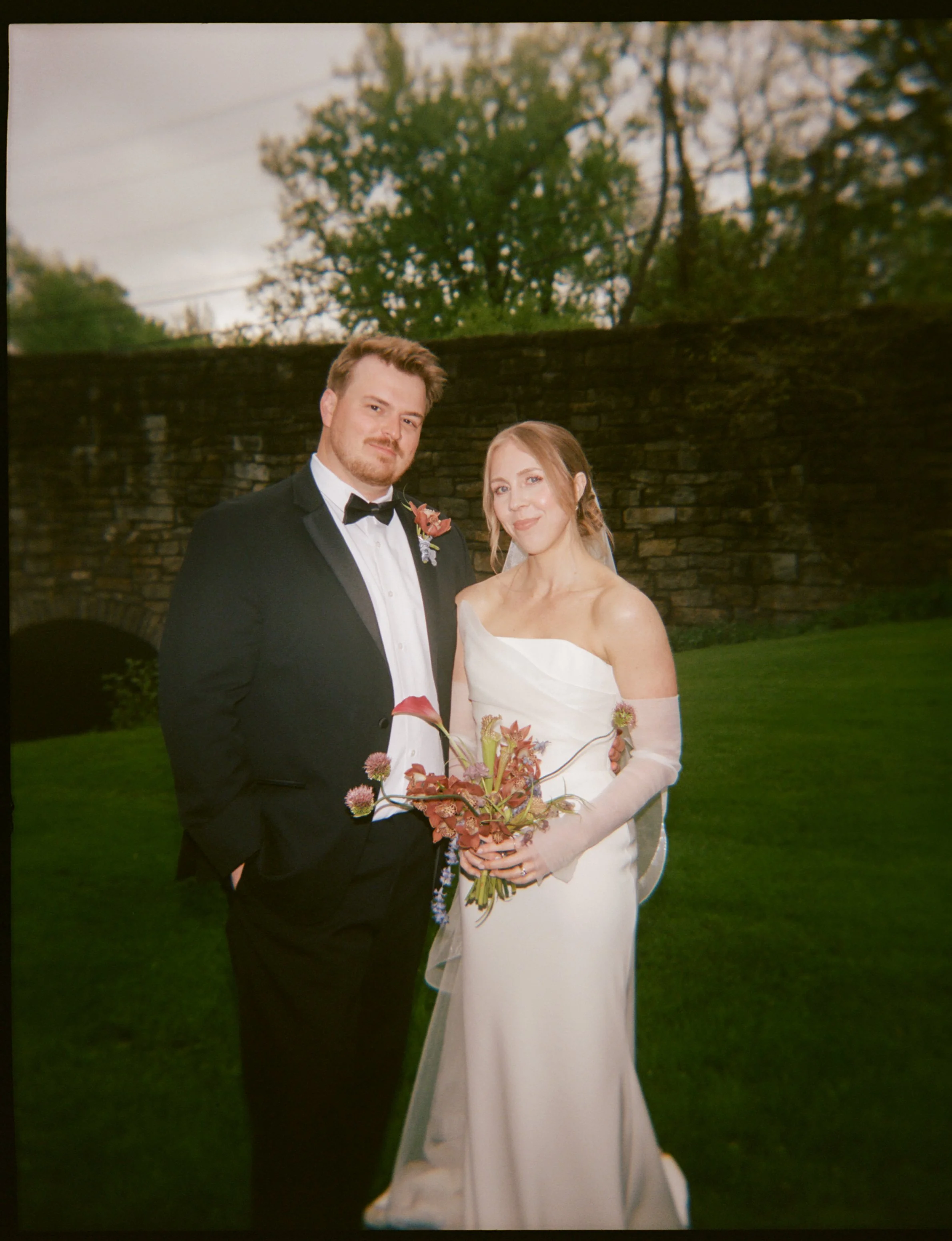 Sydney stands with husband, Josh, and  holds a whimsical petite bouquet in front of Elizabeth House in Leeds, NY at her intimate backyard micro-wedding in the Catskills. Film photography by Mikala Gallo Photography.