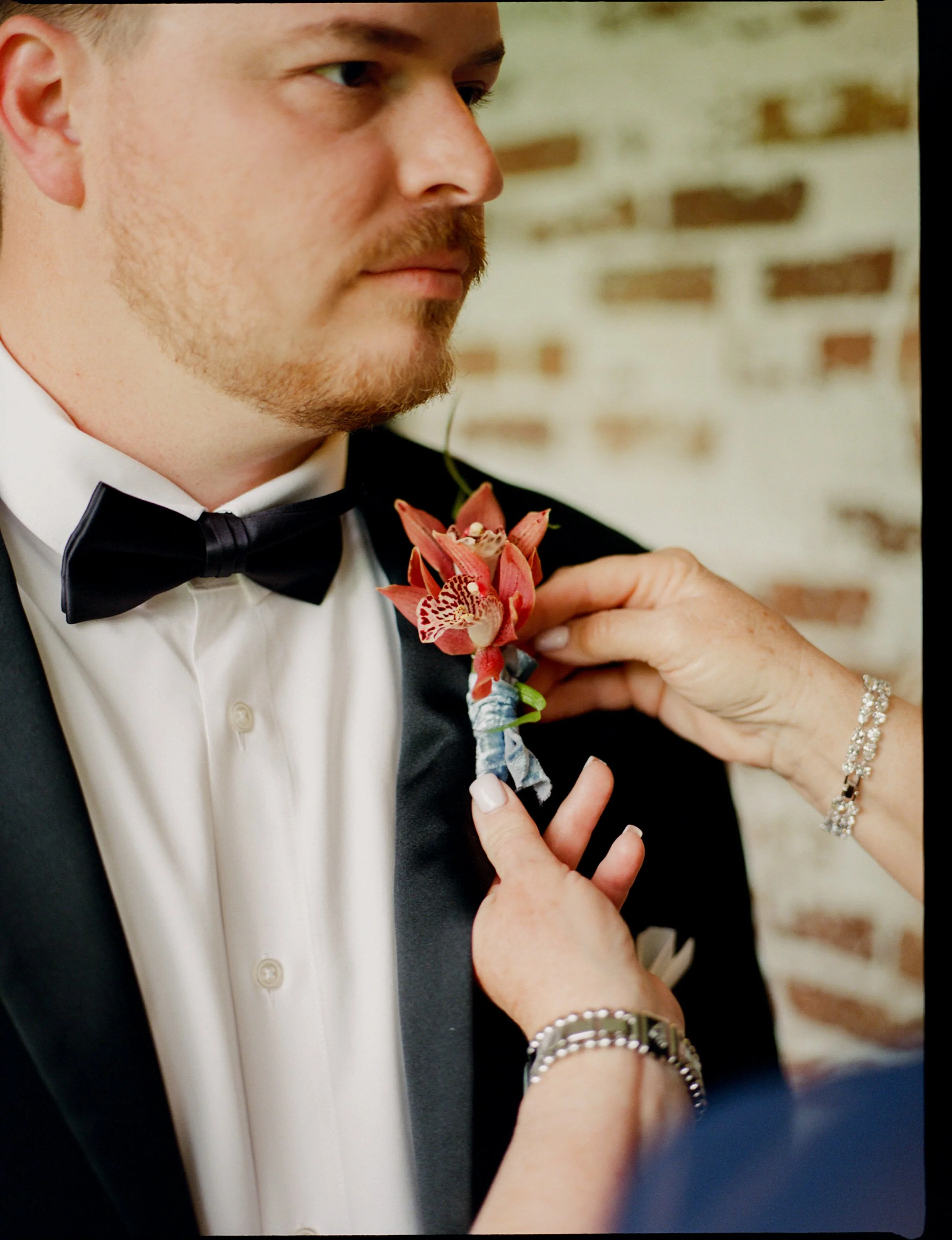 A man dressed in a tuxedo with a bow tie and white shirt, is having a boutonniere with a pink flower pinned to his lapel by a woman, during a wedding or formal event.