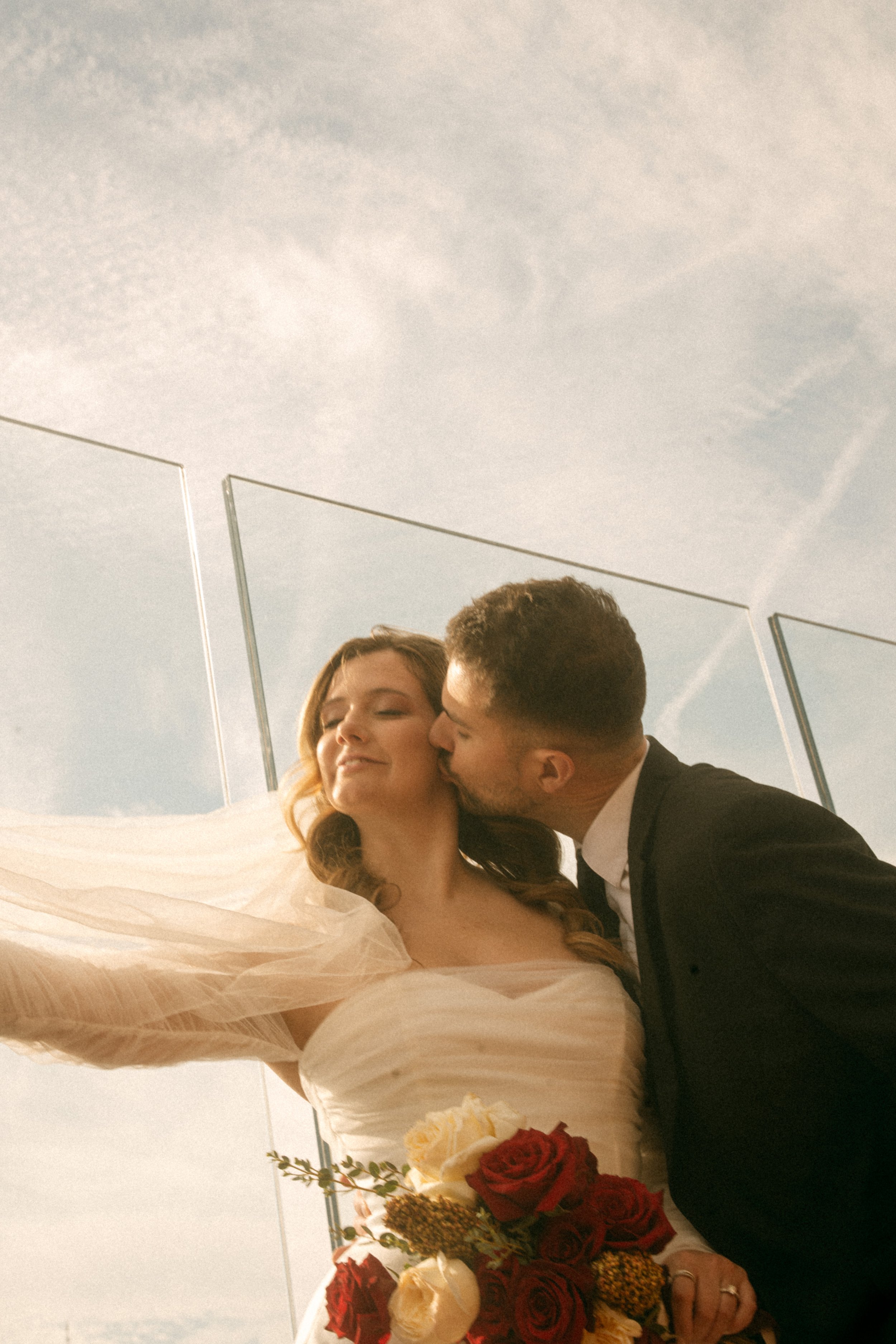 Tim & Emily kiss at "Top of The Rock."  Emily holds a gorgeous red and white classic bouquet designed by Peekskill based florist, Raving Flamingo Flowers. Photo by Brittany Dobrowski, makeup by Lemony NYC.