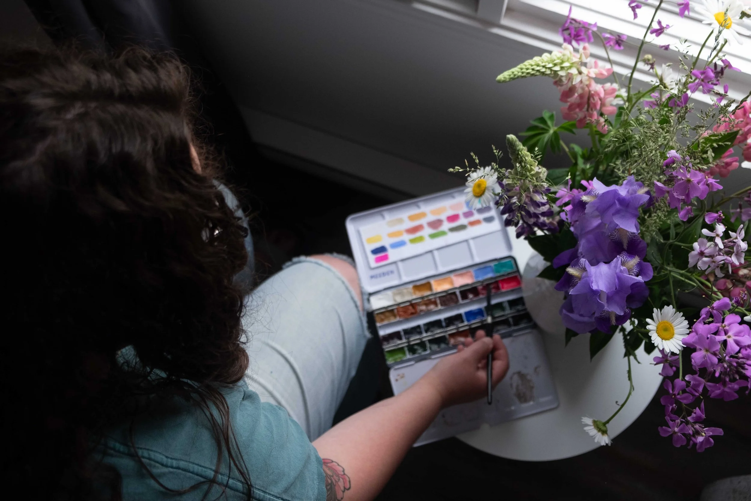 Josée Landry, artiste multidisciplinaire du Kamouraska, tenant sa palette d'aquarelles devant un bouquet de fleurs colorées dans son atelier.