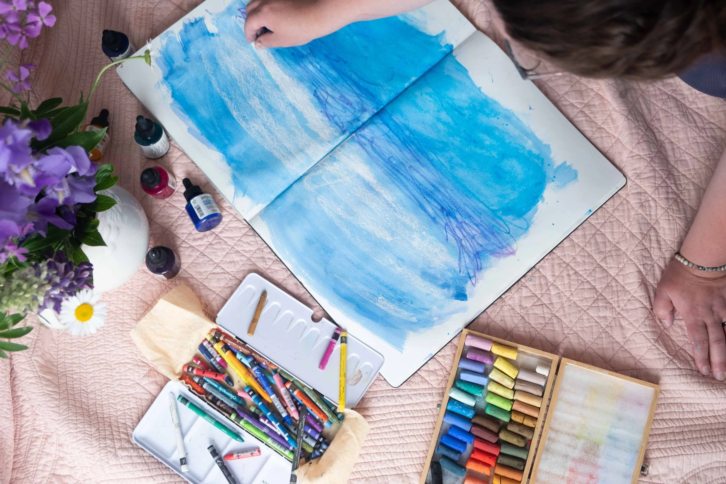 Josée Landry travaillant un paysage à l'acrylique et au pastel dans son carnet de croquis, entourée de fleurs et matériaux artistiques, atelier créatif au Kamouraska.