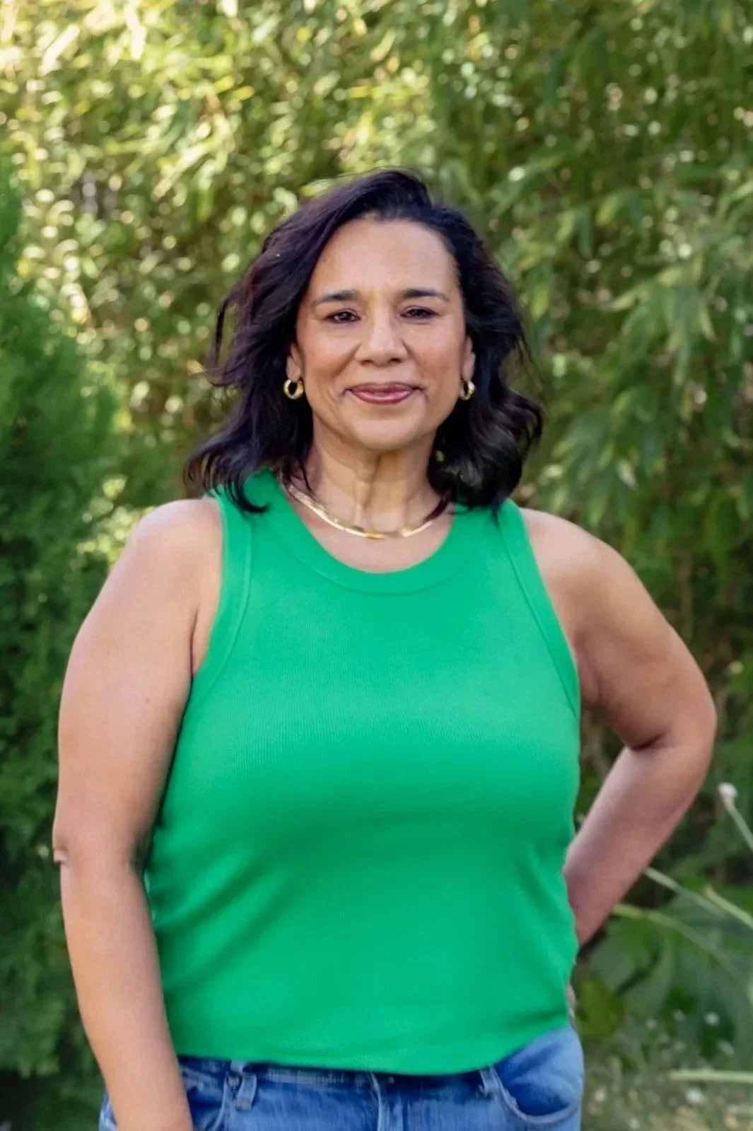 A woman with dark wavy hair, wearing a green sleeveless top, gold hoop earrings, and a gold necklace, standing outdoors amid lush green foliage.
