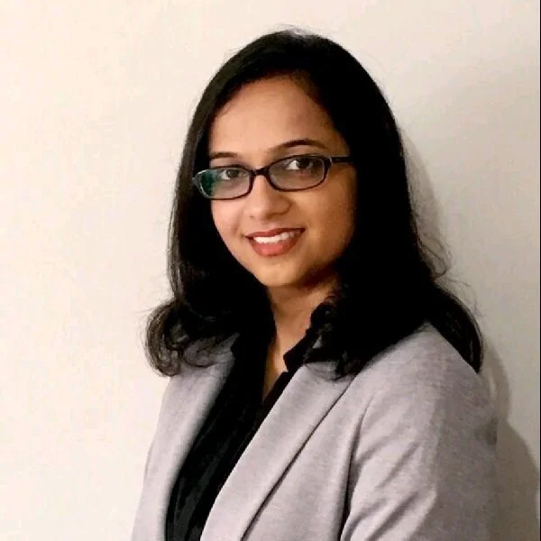 A woman with shoulder-length dark hair, wearing glasses, a gray blazer, and a black blouse, smiling at the camera against a plain white wall.