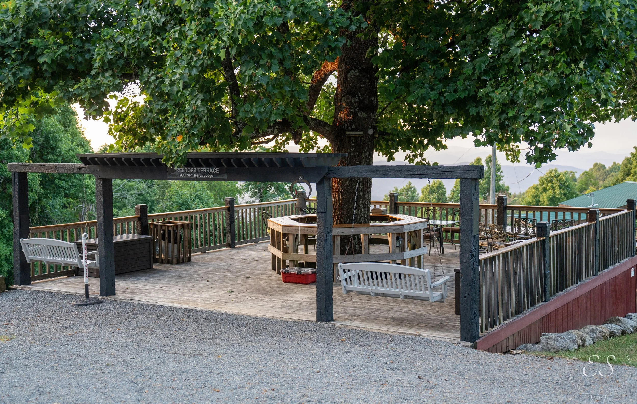 A wooden outdoor deck with a large tree in the center, surrounded by a black railing. The deck has benches, tables, chairs, and a swing hanging from a wooden frame, overlooking green trees and a distant mountain landscape.
