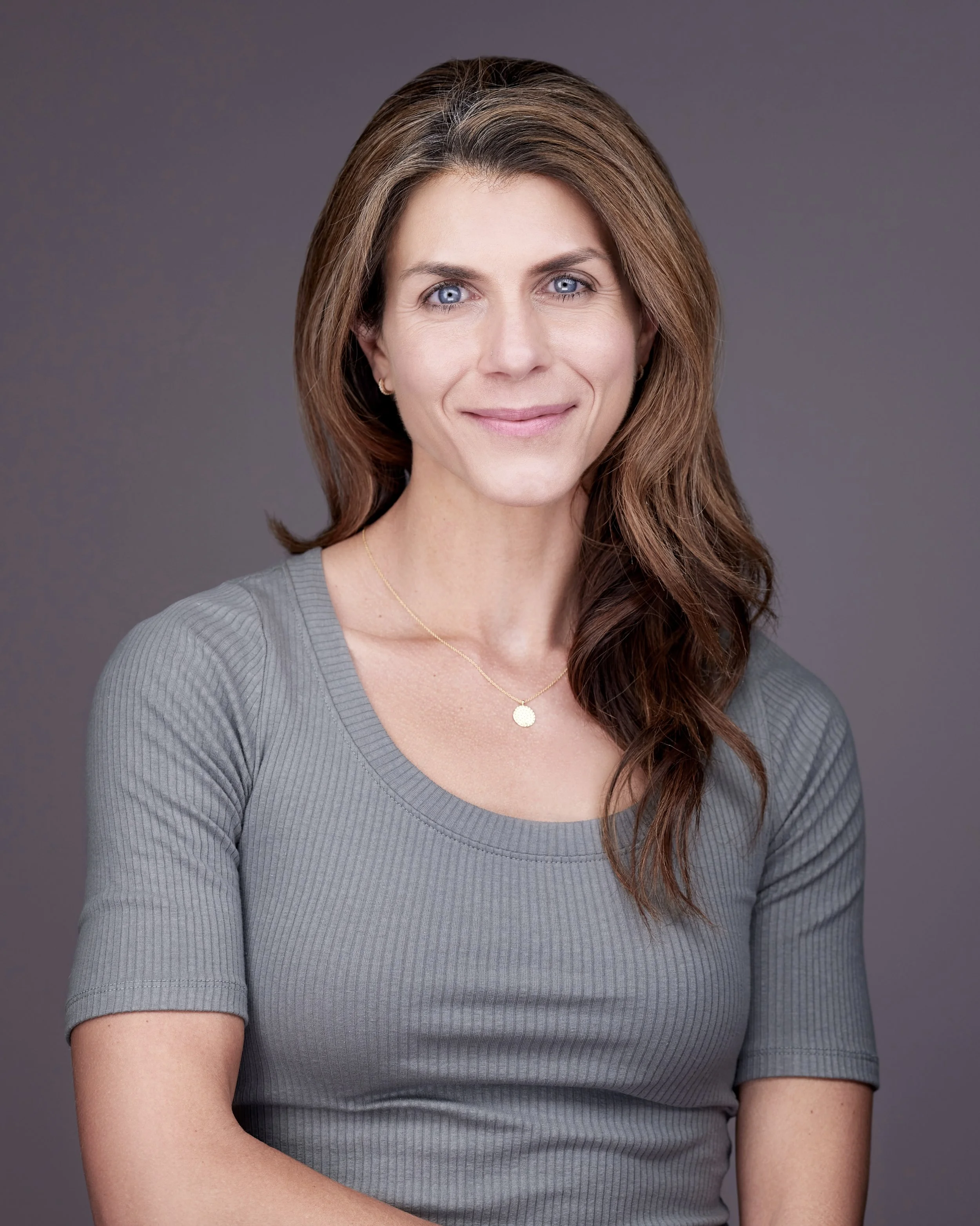 Portrait of a woman with light skin, long wavy brown hair, blue eyes, wearing a gray top and a delicate gold necklace, smiling softly against a plain gray background.