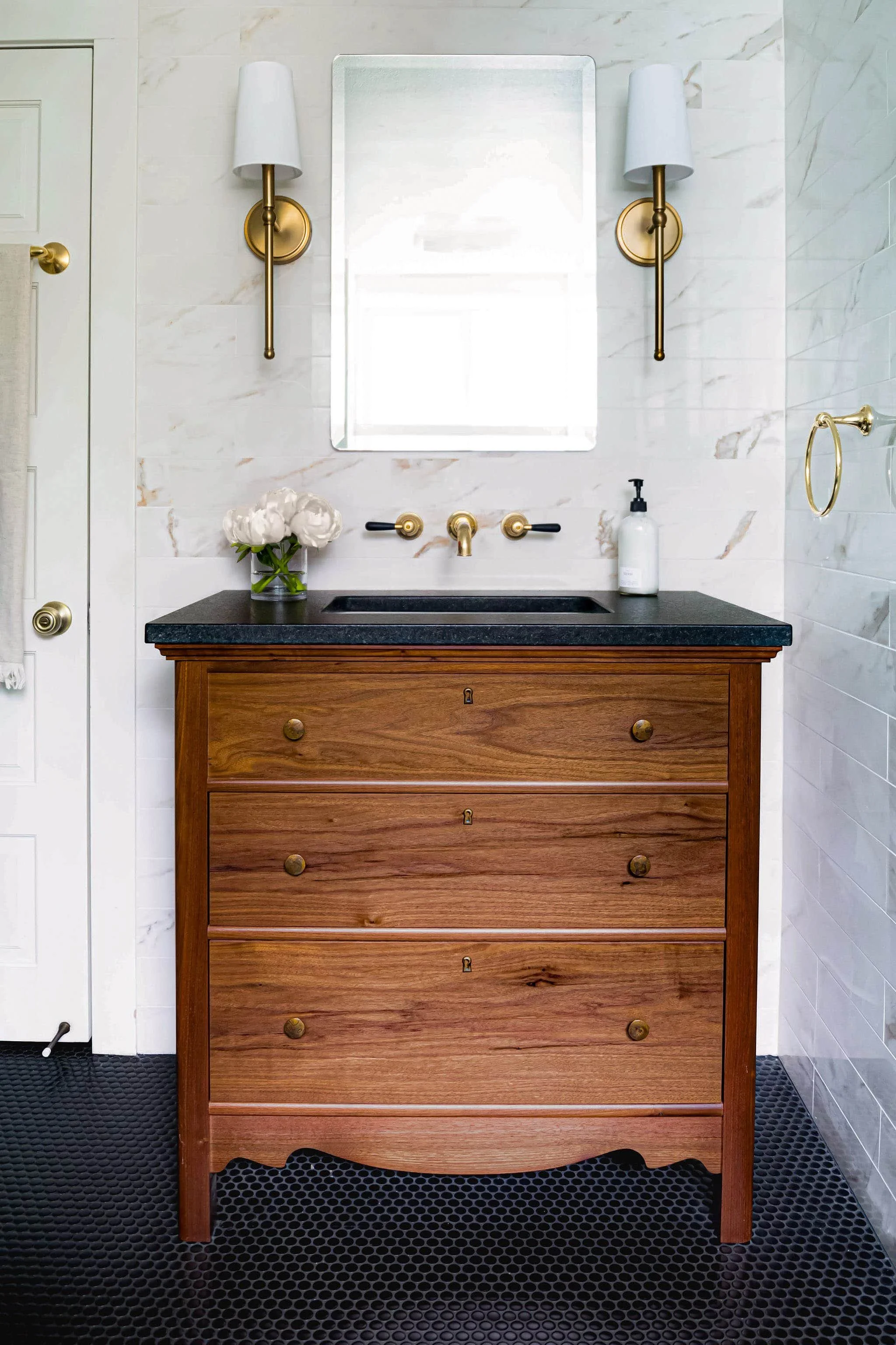 Bathroom with custom wooden vanity, black countertop, gold fixtures, round mirror, wall-mounted lamps, and white tiles.