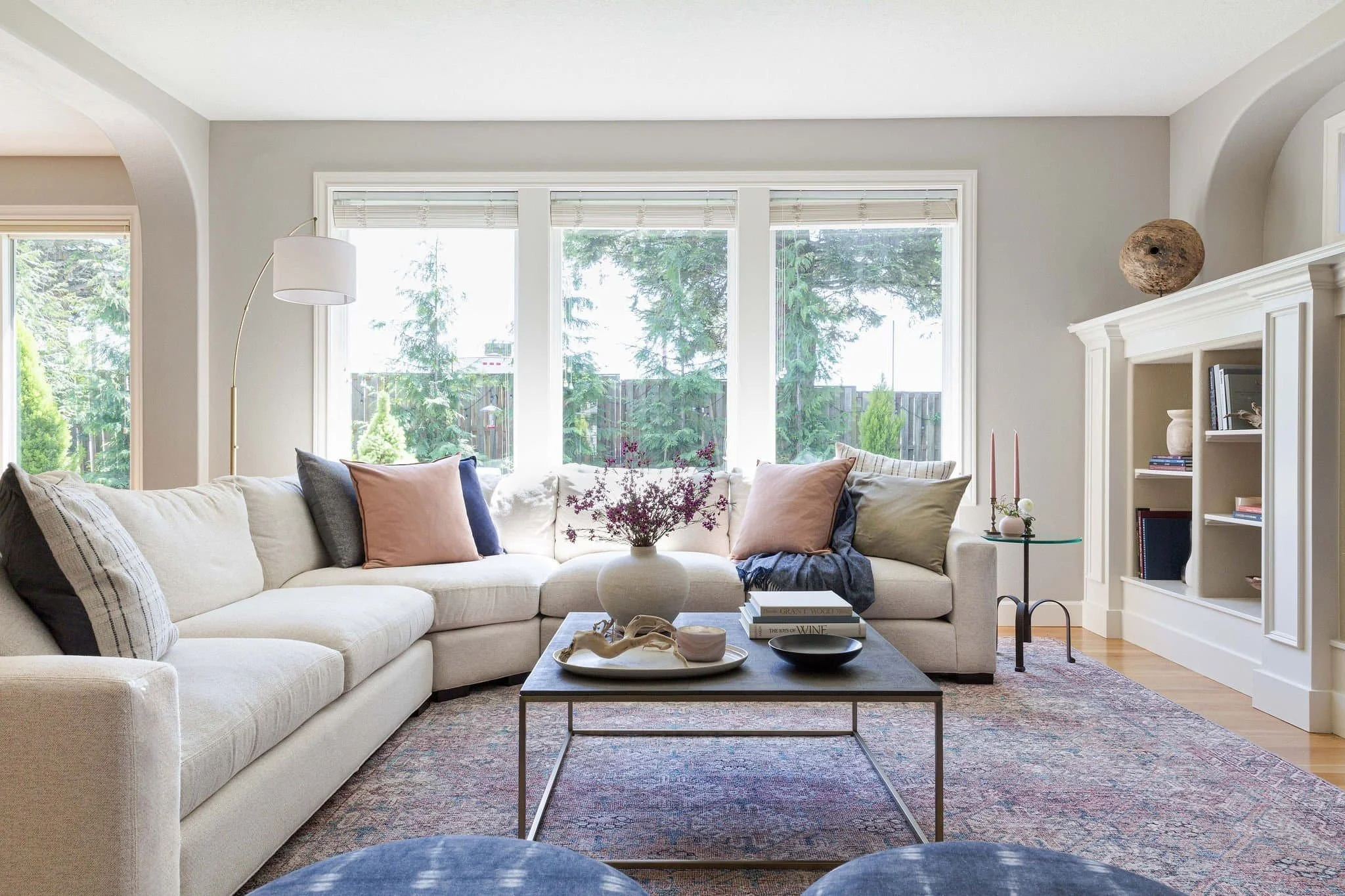 Bright living room with a beige sectional sofa, colorful pillows, a wooden coffee table with decorative objects, a white vase with flowers, and large windows showing greenery outside.