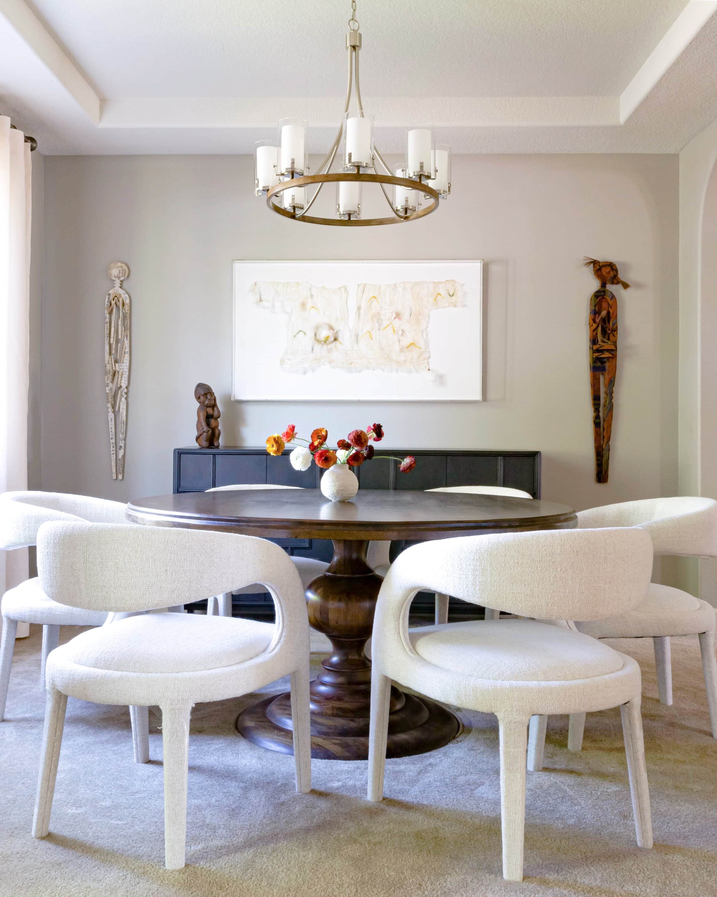 A dining room with a round wooden table and white upholstered chairs. A chandelier hangs above the table. The walls feature abstract art and wooden sculptures. A small vase with flowers is on the table.