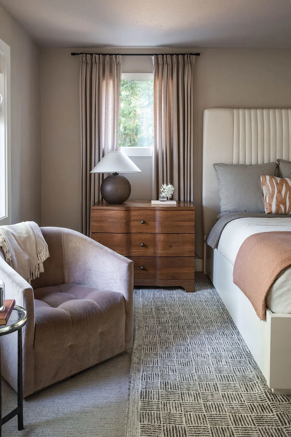 Primary bedroom corner view with a soft upholstered chair, wood dresser and table lamp, layered bedding, and neutral drapery framing the window.