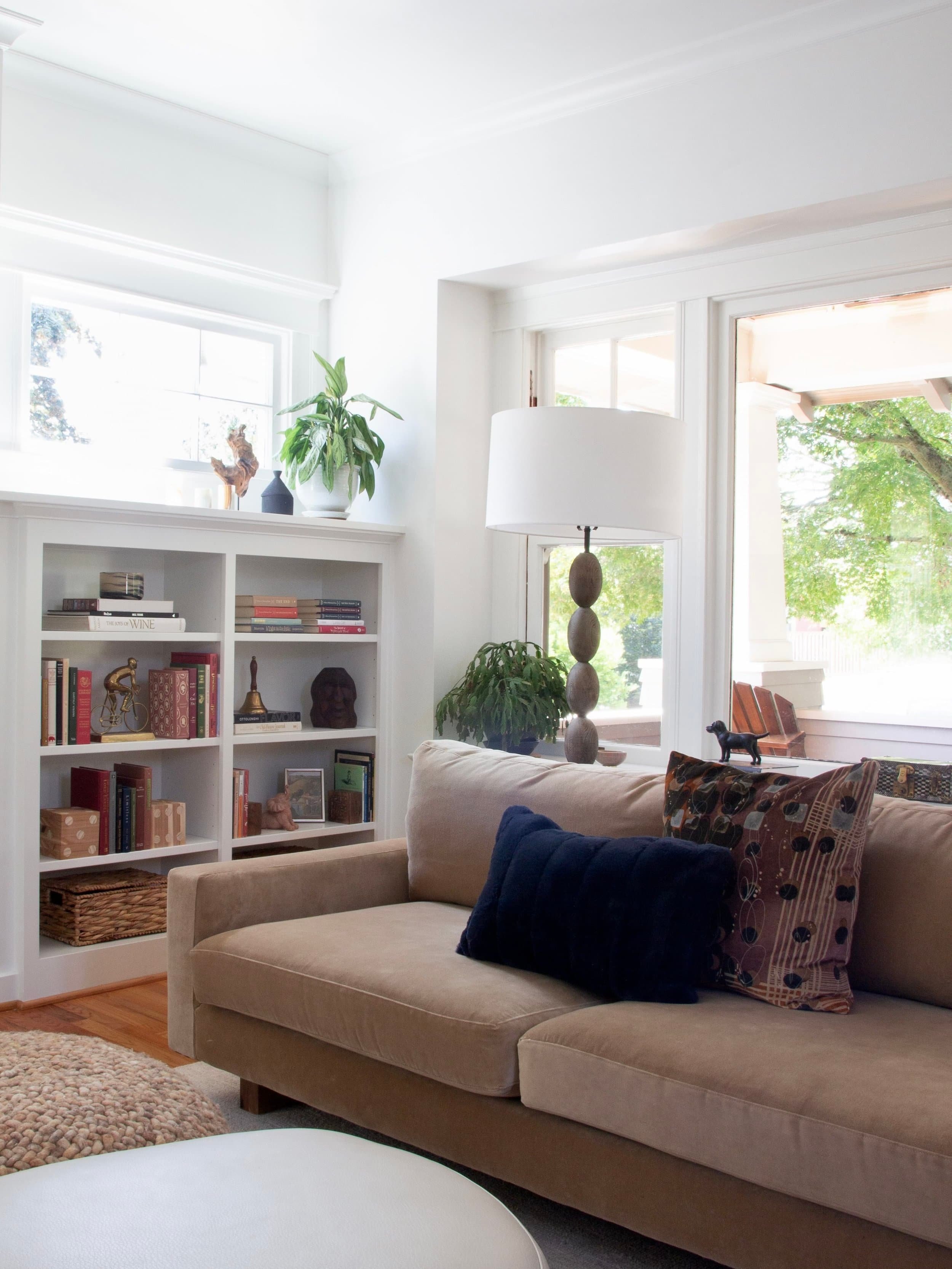 Living room with beige sofa, blue and patterned cushions, white bookshelf with books and decor, indoor plant, modern table lamp, round woven rug, and large windows with outdoor view.
