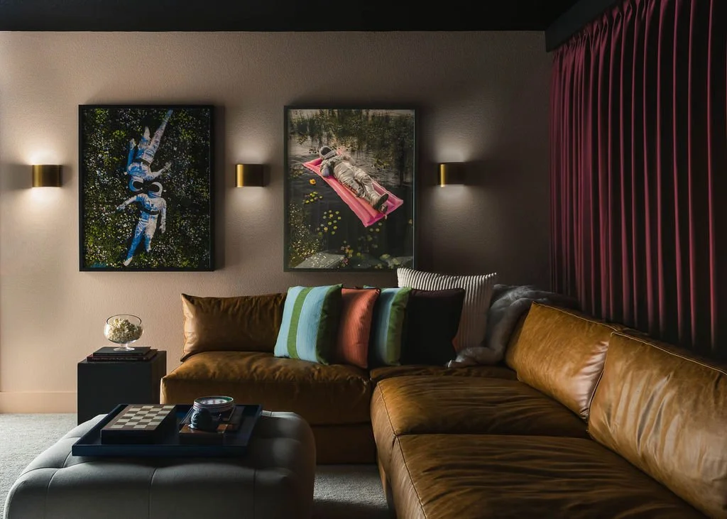 Media room with a caramel leather sectional sofa, layered striped and solid pillows, two framed art pieces above the seating, warm brass wall sconces, a dark coffee table, and deep plum curtains creating a cozy, moody atmosphere.