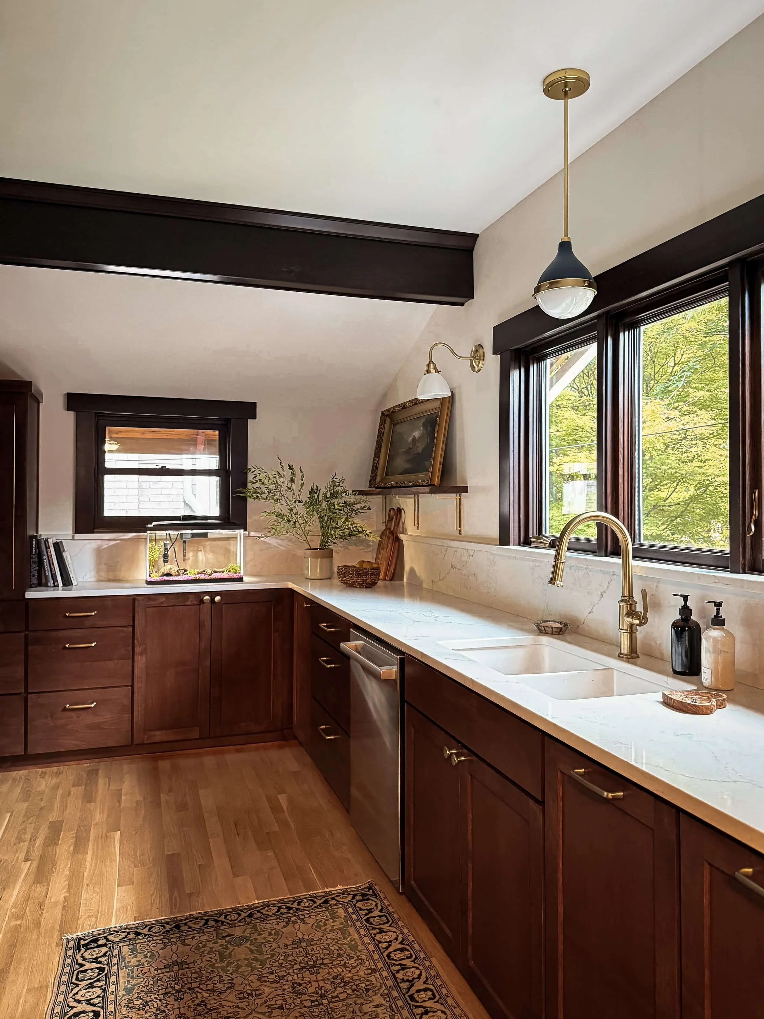 Kitchen with dark wood cabinets, white marble countertops, gold fixtures, and large windows showing green trees outside.