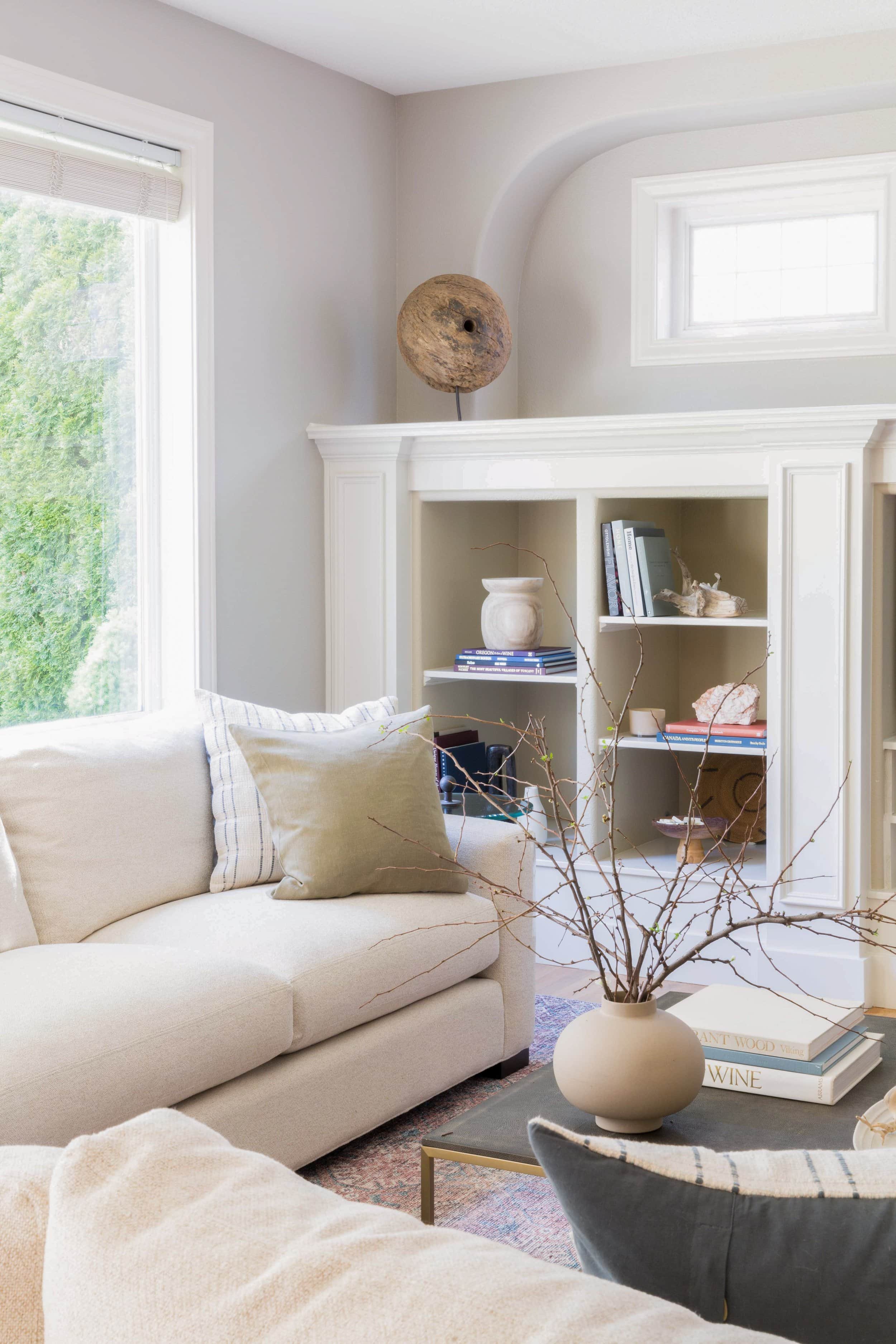 Modern living room with beige sofa, decorative pillows, a coffee table with a round vase holding branches, and a bookshelf with books and decorative items. A large window provides natural light.