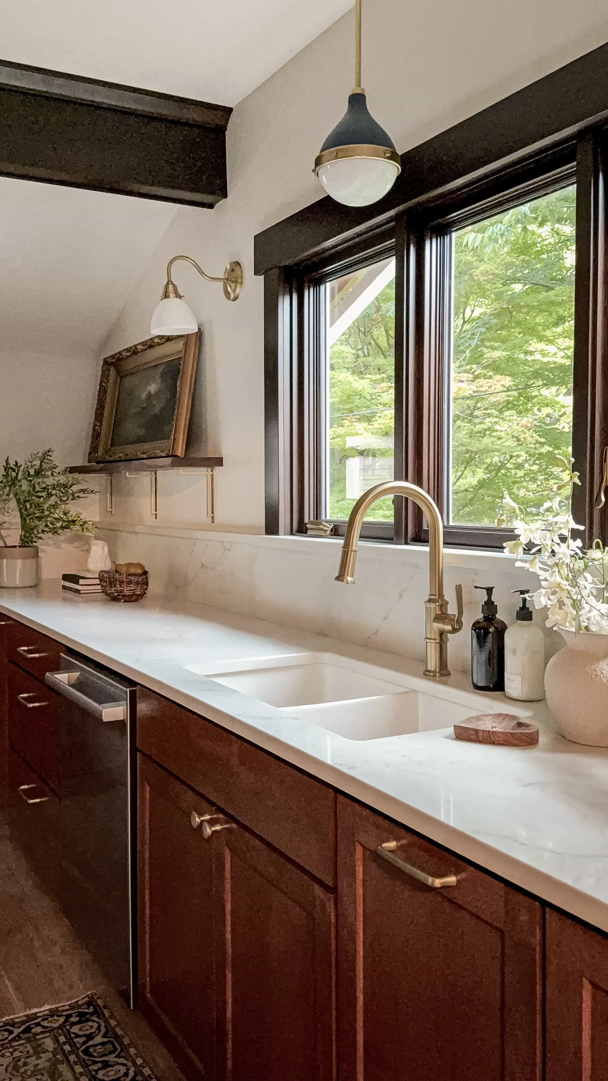 Kitchen with white marble countertops, wooden cabinets, a gold faucet, a window with trees outside, and decorative items like a plant, framed picture, and soap dispensers.