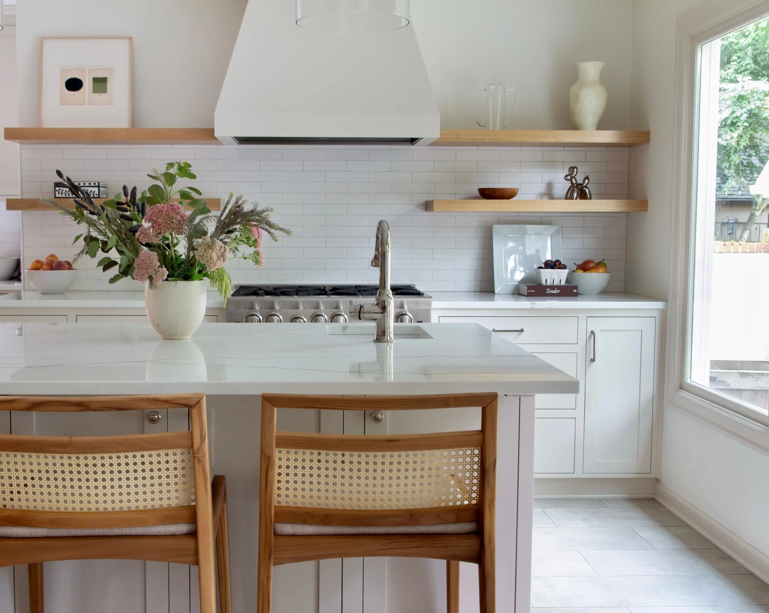 White modern kitchen with wooden accents, a white countertop with a faucet, a vase with pink flowers, wooden chairs, a stove, open wooden shelves with decorative items, a bowl of fruit, and a large window with a view of trees outside.