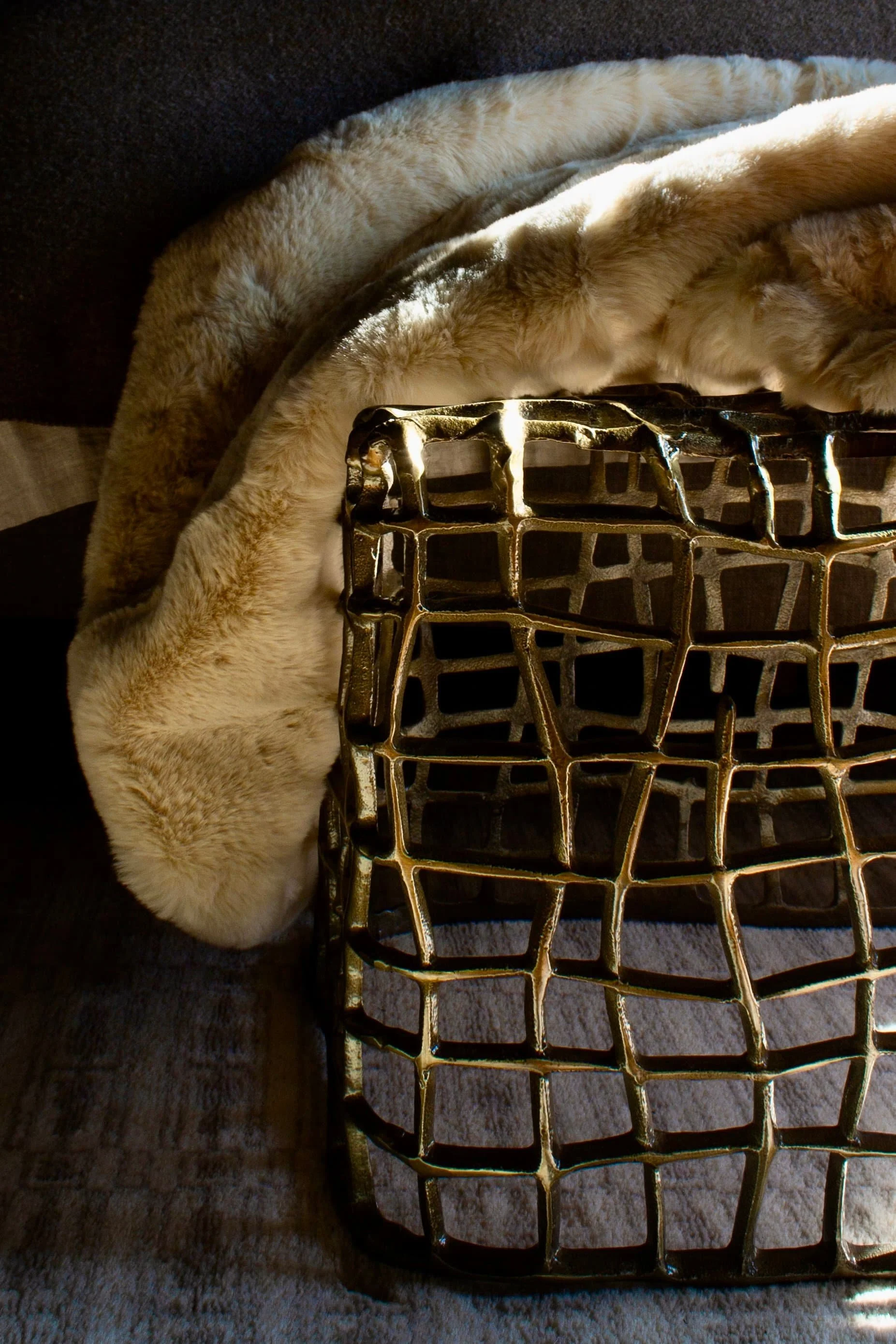 Abstract gold metal grid sculpture on a soft beige fur blanket over dark background.