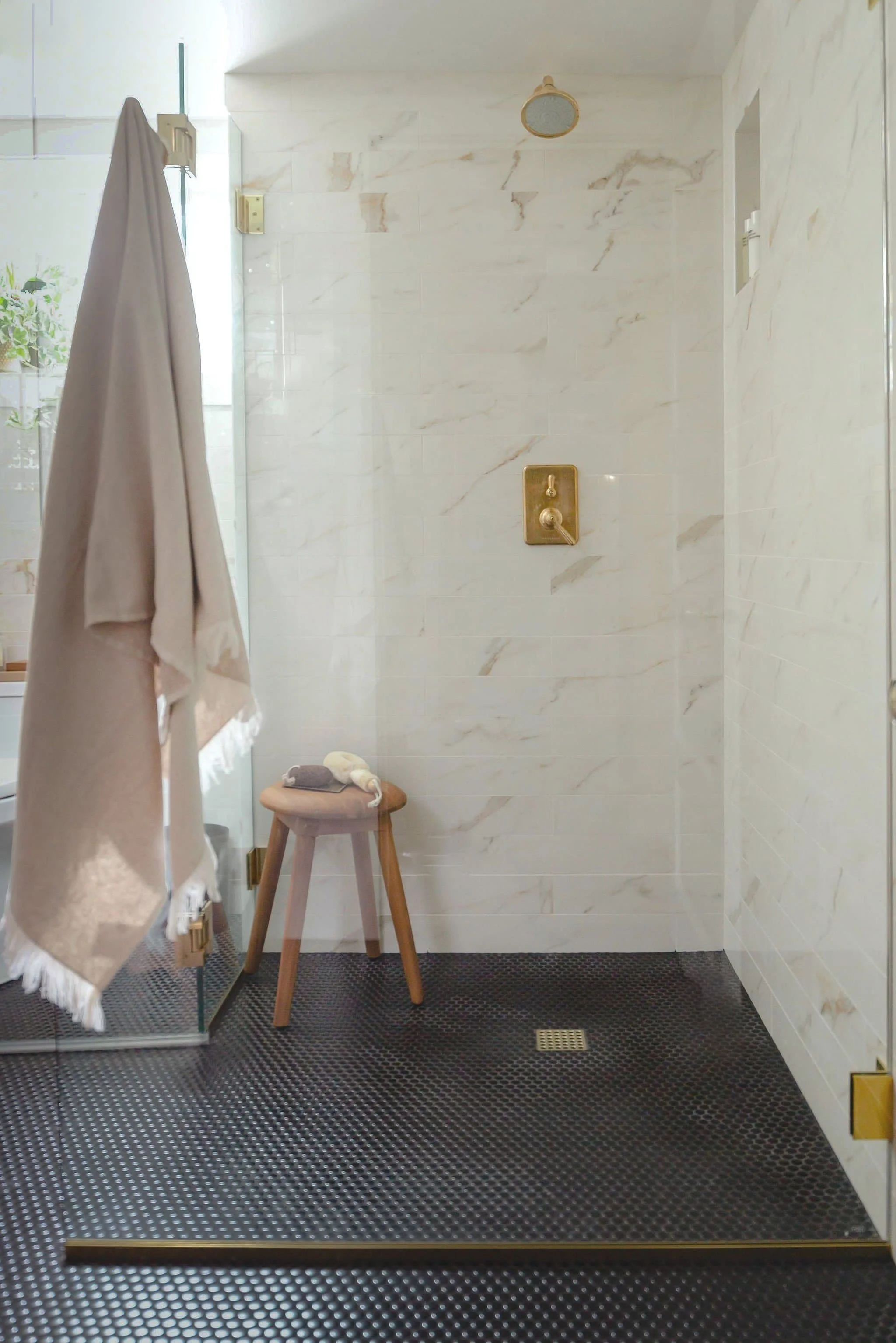 Modern bathroom with a glass shower, white marble tiles, wooden stool, and hanging towel.