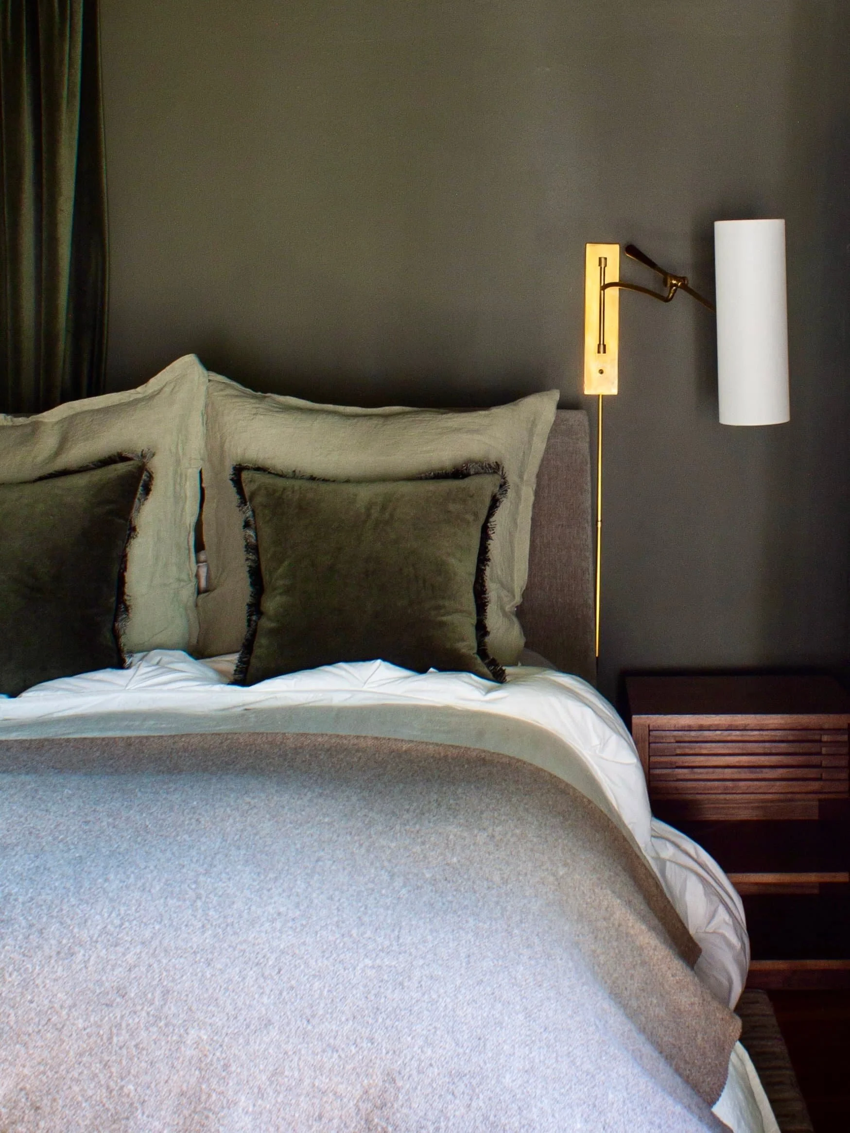 Modern bedroom interior with a bed featuring dark green pillows and a gray blanket, a bedside table, and a brass wall-mounted lamp against a dark green wall.